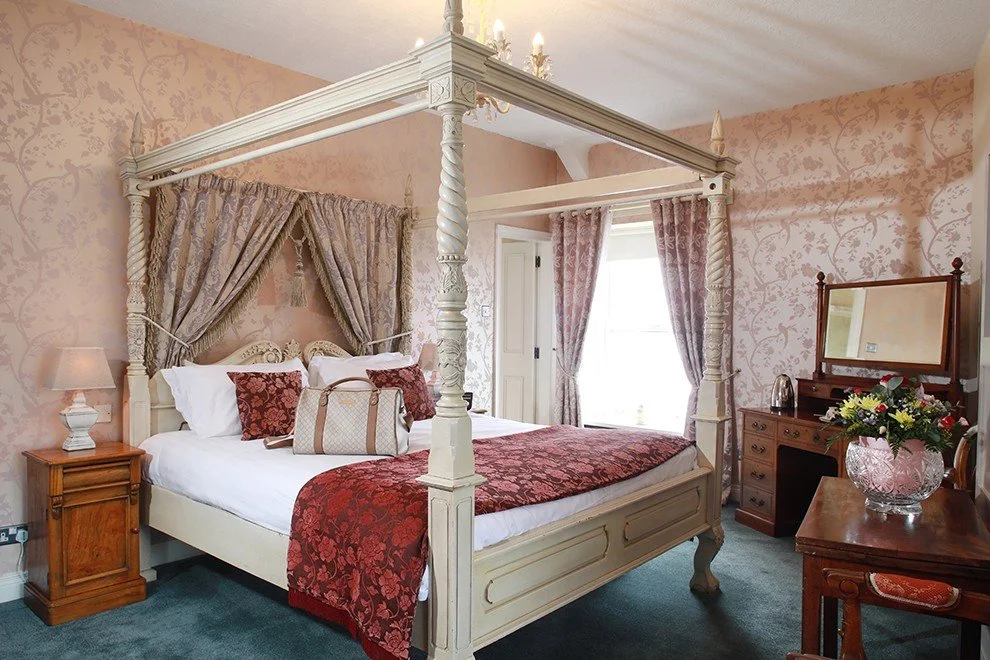 A bedroom with a cream-colored four-poster bed with ornate carved posts, draped with patterned curtains, and topped with a chandelier. There are red and white pillows, a patterned bedspread, and a handbag on the bed. The room has pink wallpaper with floral patterns, a wooden nightstand with a lamp, a wooden dresser with a mirror, and a vase of flowers on a table. Natural light enters through a window with curtains.