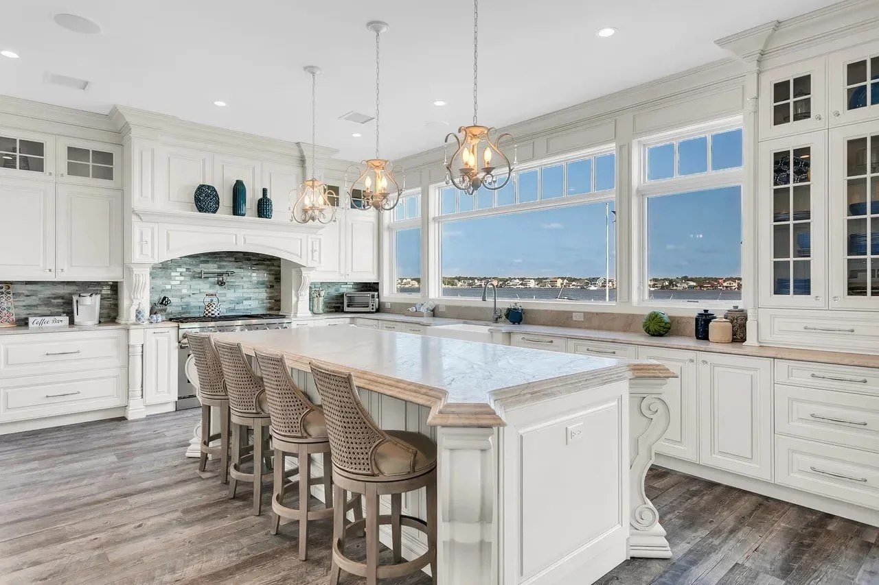 Bright, spacious kitchen with white cabinetry, large windows overlooking water, marble countertop island, wooden bar stools, and modern light fixtures.