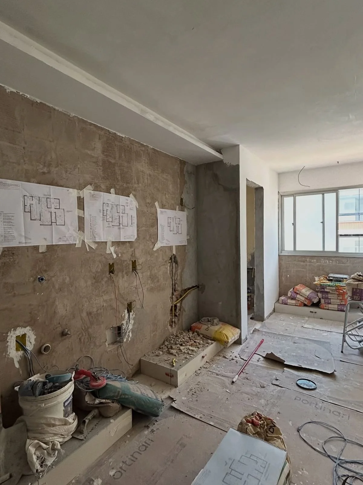 Interior view of a room under renovation with construction materials, wiring, and plans taped to the wall.