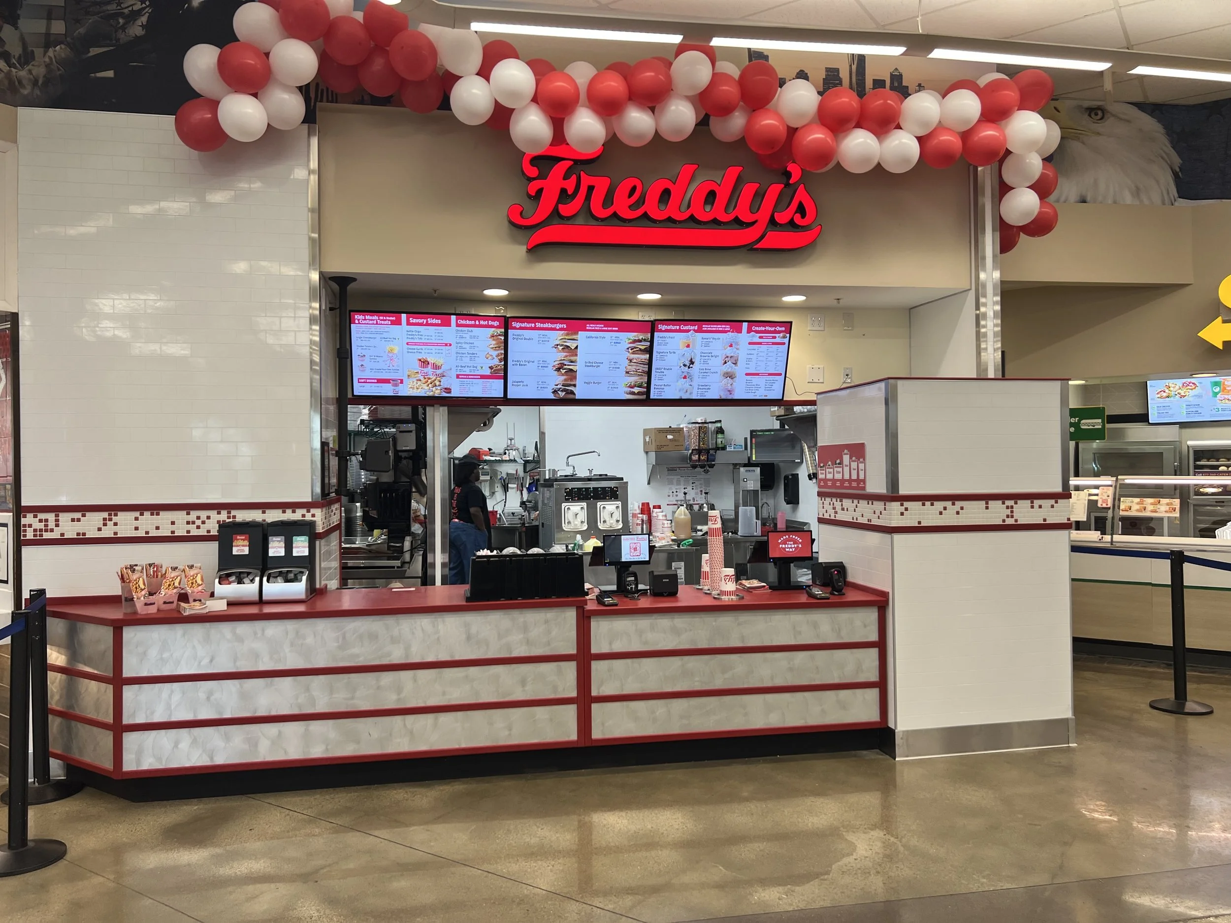 Freddy's fast-food restaurant counter with a red and white menu board, balloons decorating the area, and a worker behind the counter.