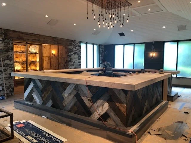 Interior of a room with a large wooden pool table, a man working on it, a hanging light fixture, and large windows with frosted glass.