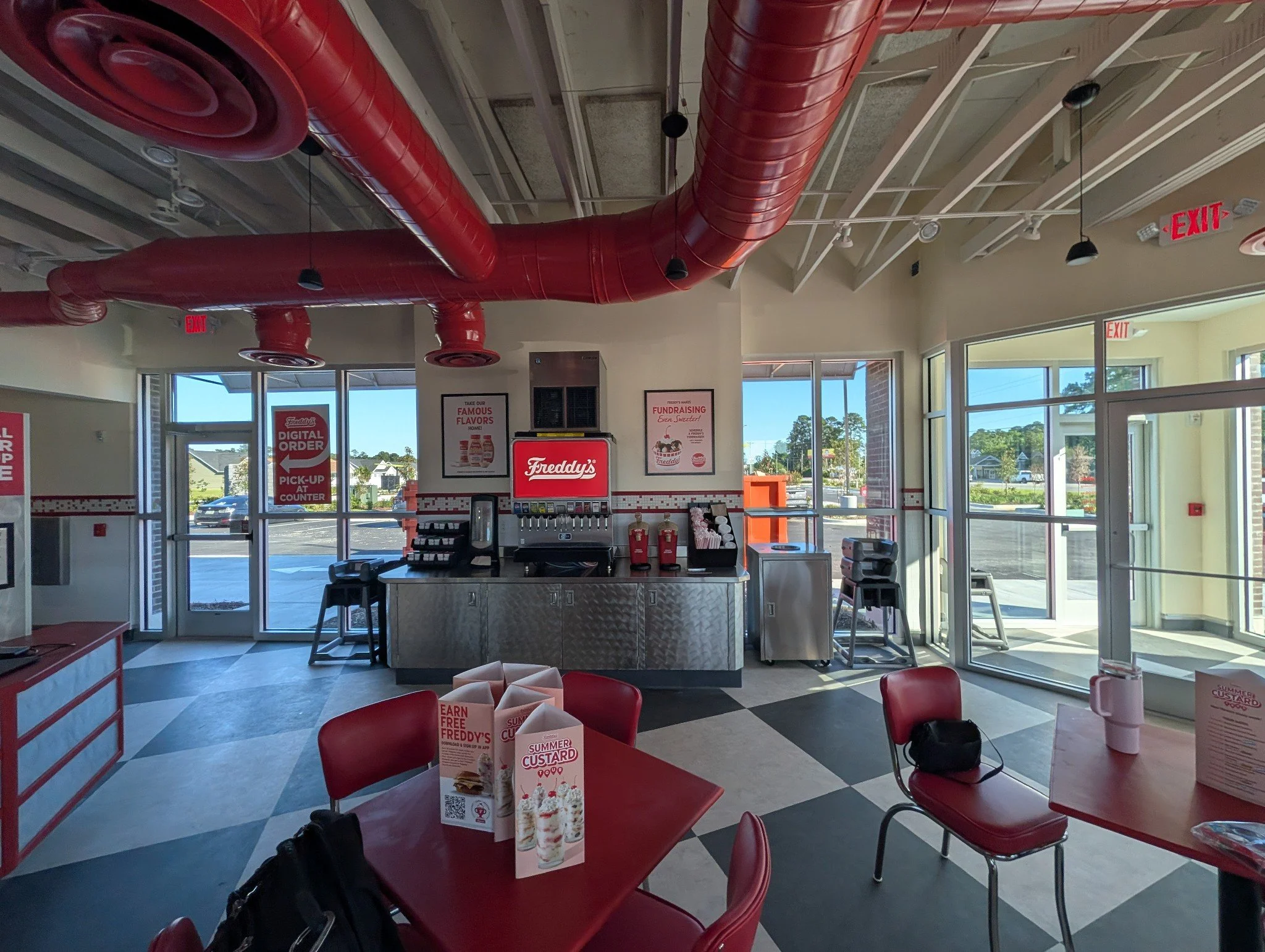 Interior of a fast food restaurant with red pipes on the ceiling, red chairs, and promotional menus on the tables, with windows showing a sunny day outside.