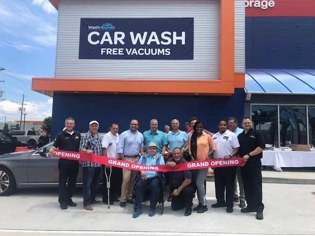 Group of 12 people at grand opening of a car wash business holding a red ribbon with 'Grand Opening' written on it, in front of a sign that says 'CAR WASH FREE VACUUMS'