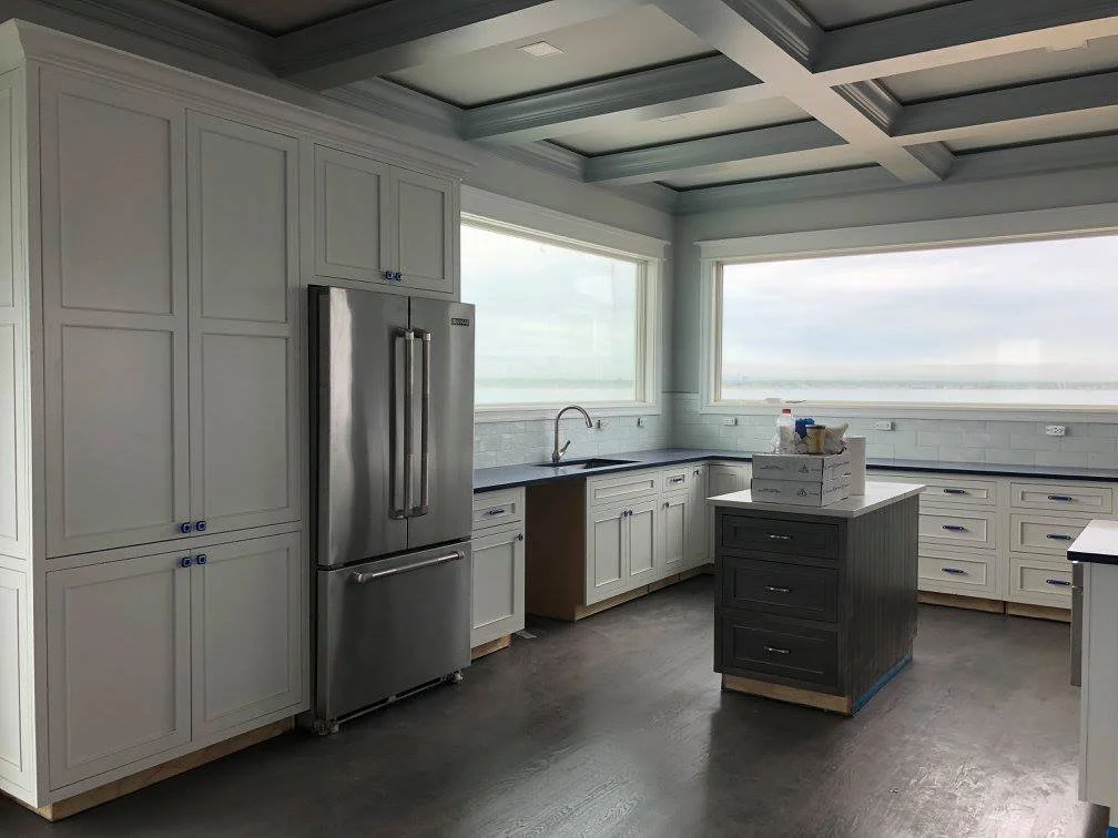 A modern kitchen with white cabinets, a stainless steel refrigerator, and large windows showing a view of the outdoors