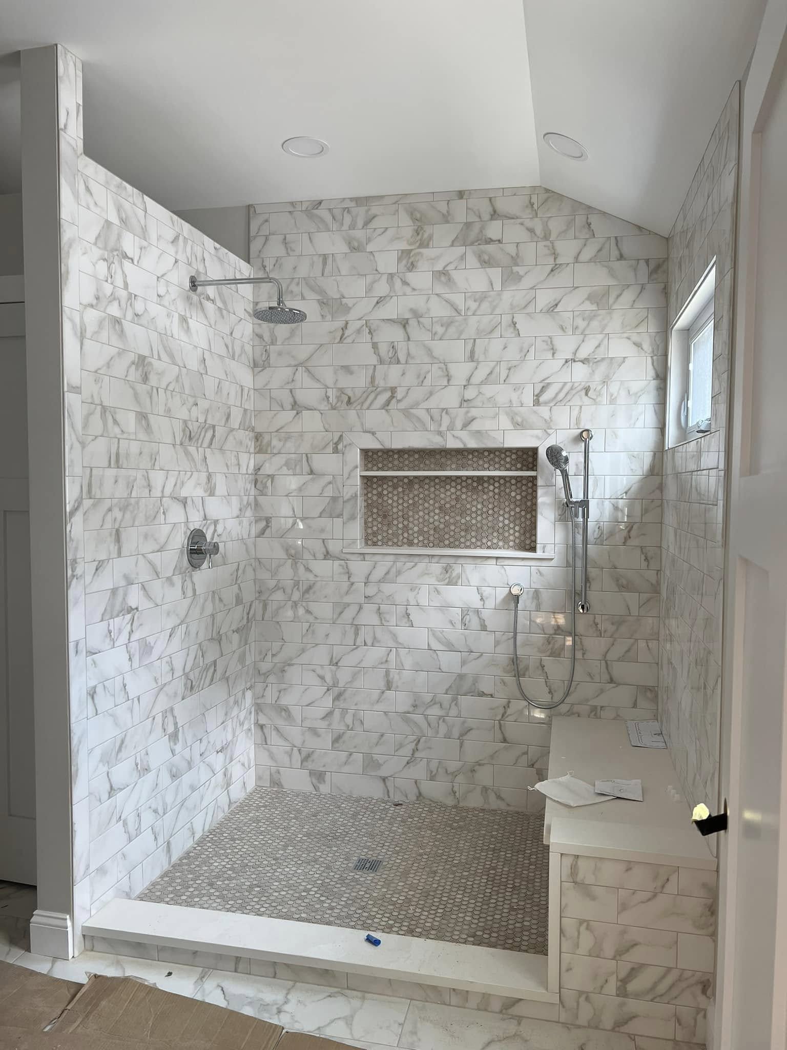 A modern walk-in shower with white marble tile walls, a rainfall showerhead, a handheld showerhead, a small window, a built-in niche with hexagonal tile, and a textured shower floor. Part of a white marble countertop with a small paper item is visibl