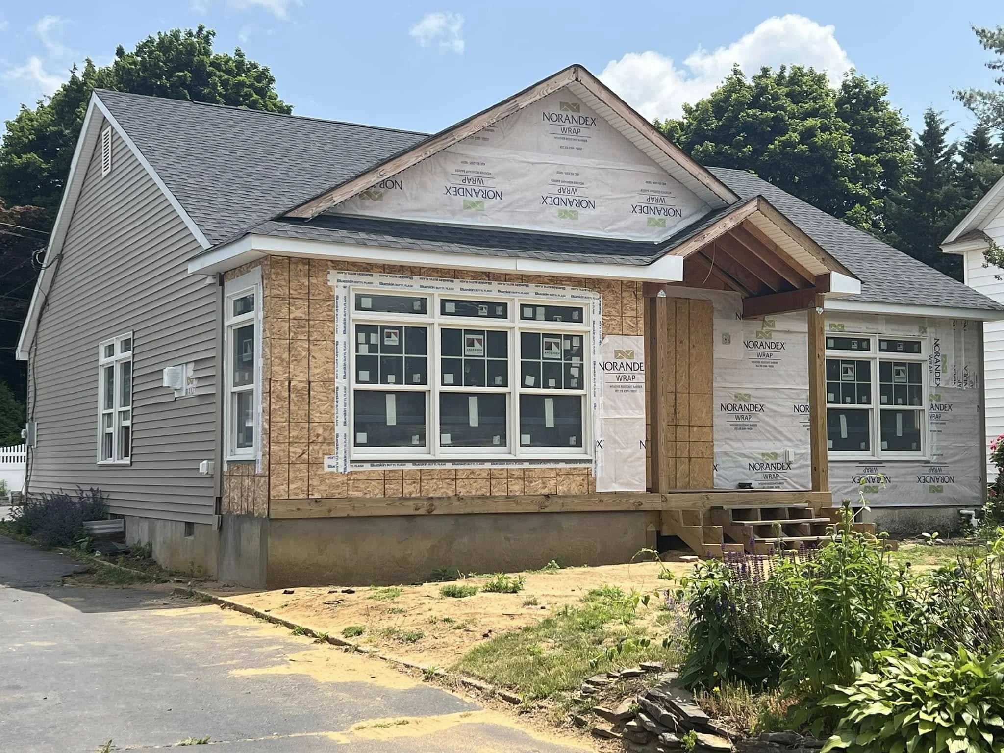 Side view of a house under construction with partially installed siding and roofing, surrounded by green trees and a clear blue sky.