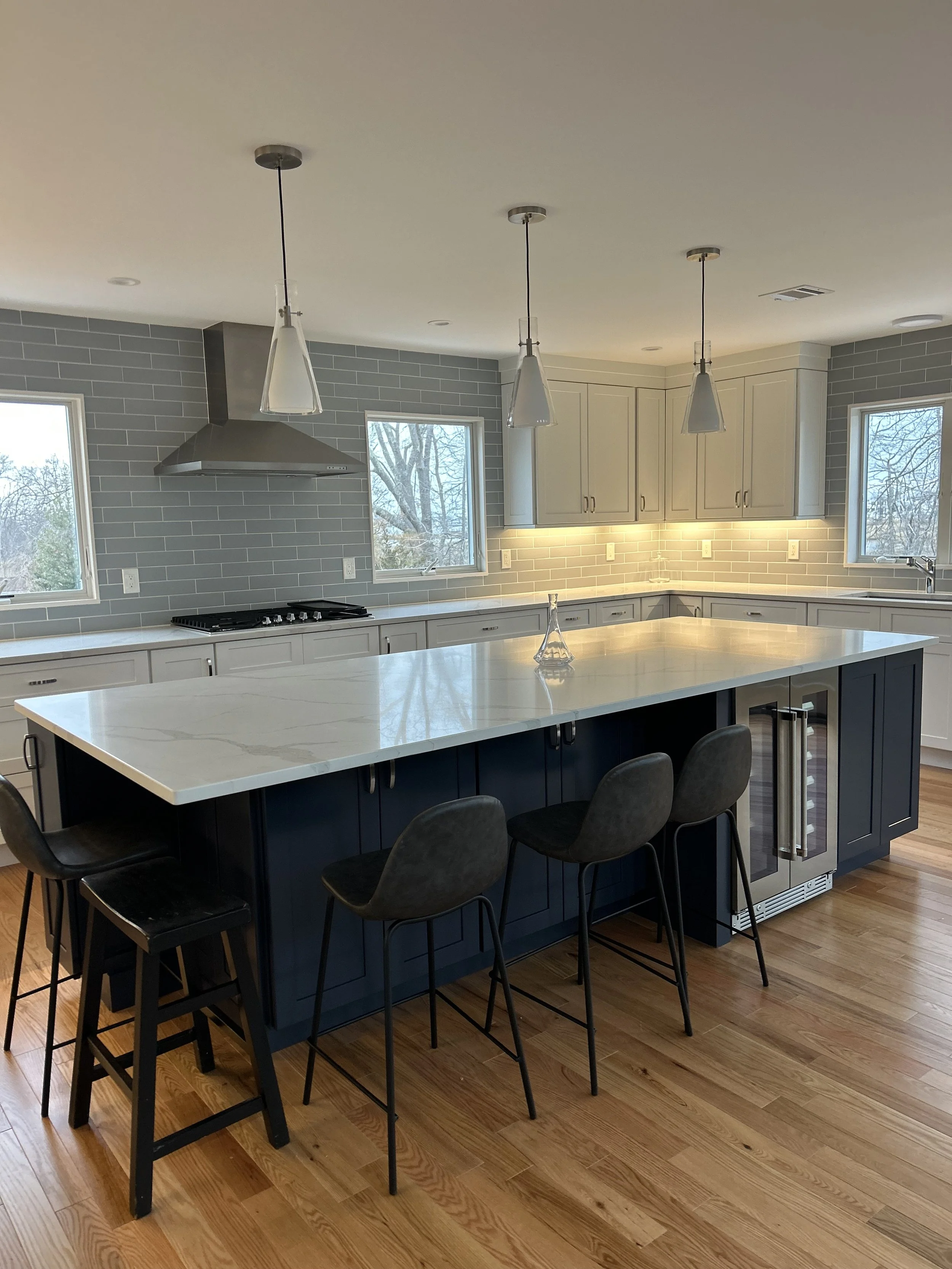 Modern kitchen with white countertops, navy blue cabinets, gray tile backsplash, pendant lights, wooden flooring, and large windows, with chairs around a central island.
