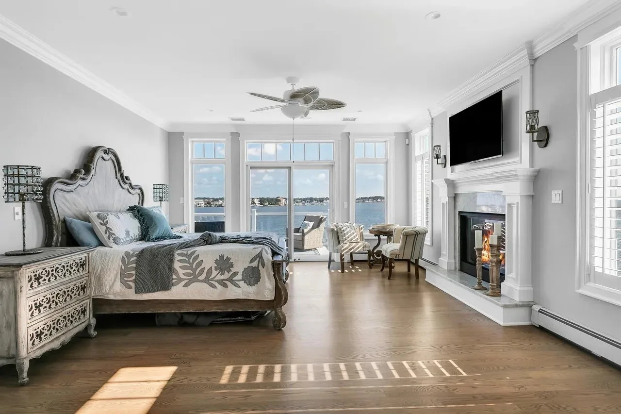 Living room with large windows overlooking water, wooden floor, fireplace, TV, bed, and seating area.