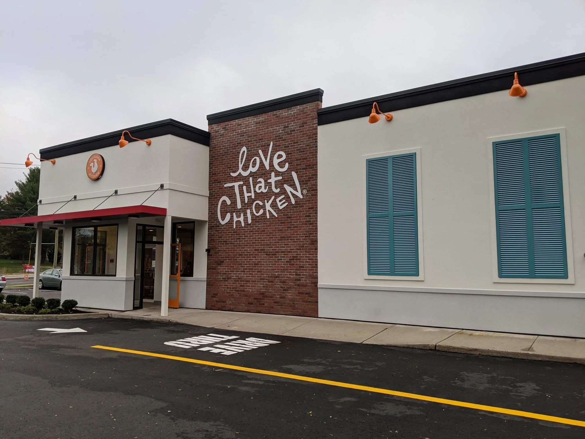 Exterior view of a Louisiana Kitchen restaurant with a white and brick facade, blue shutters, and orange lighting fixtures, located near a parking lot and street.