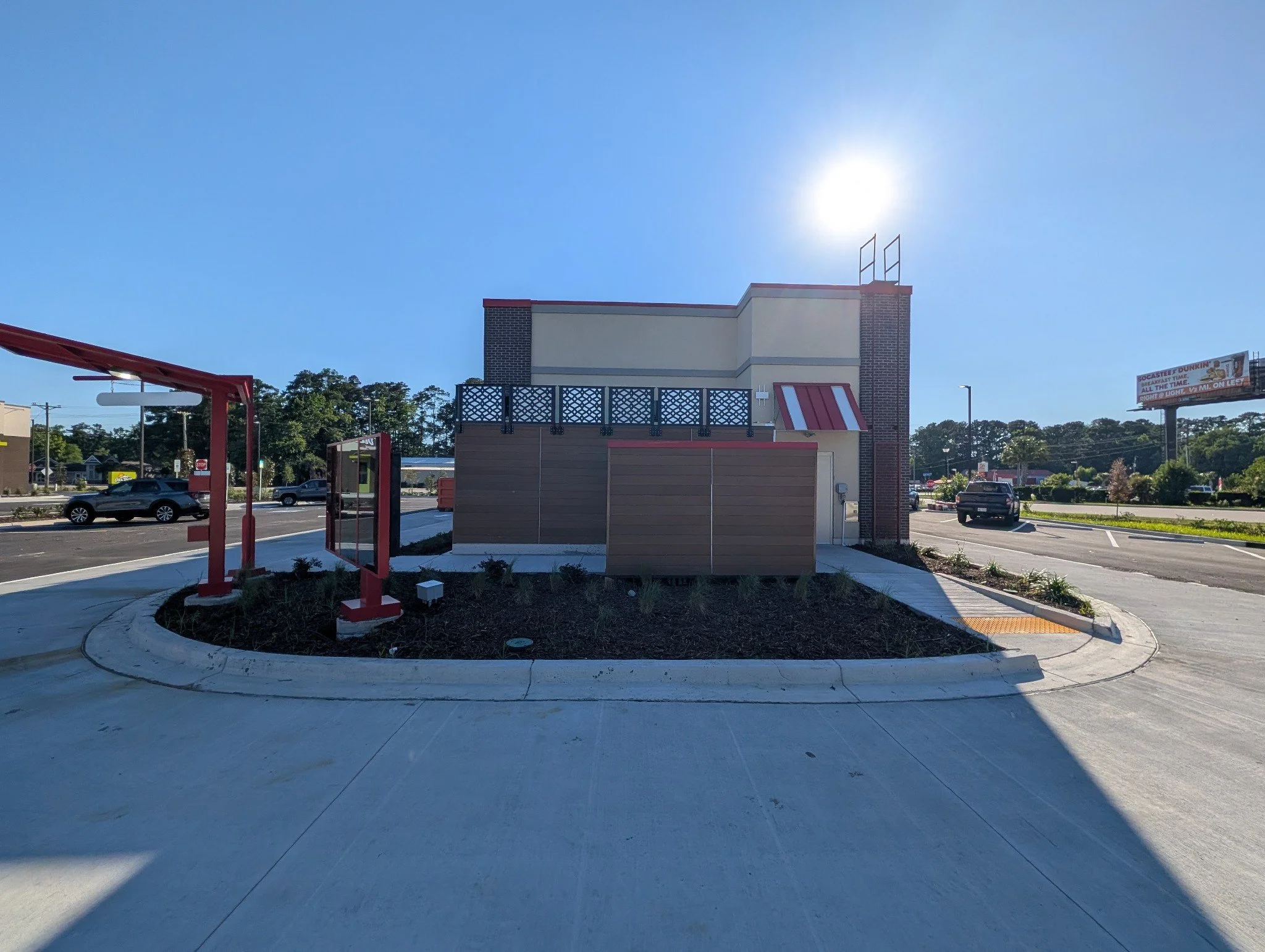A fast food restaurant parking lot with a small building, parking spaces, cars, and a billboard. The sky is clear with the sun shining brightly.