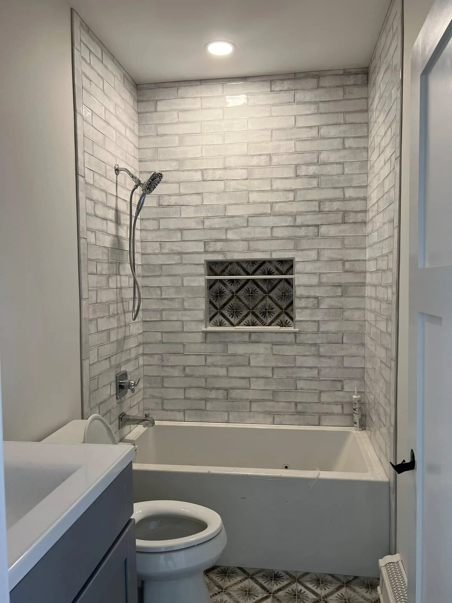 A bathroom with tiled walls, a shower with a showerhead, a toilet, a vanity with a sink, and a decorative tiled niche in the wall. The floor has patterned tiles, and there is a window on the right.