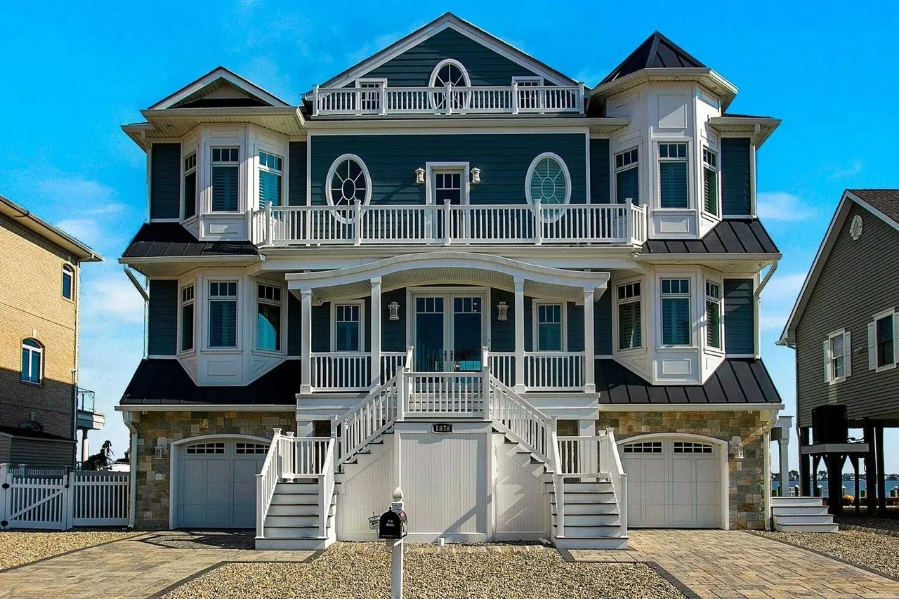 Multi-story blue house with white trim, multiple balconies, bay windows, and a staircase leading to the front door, with neighboring houses on sides and a blue sky above.