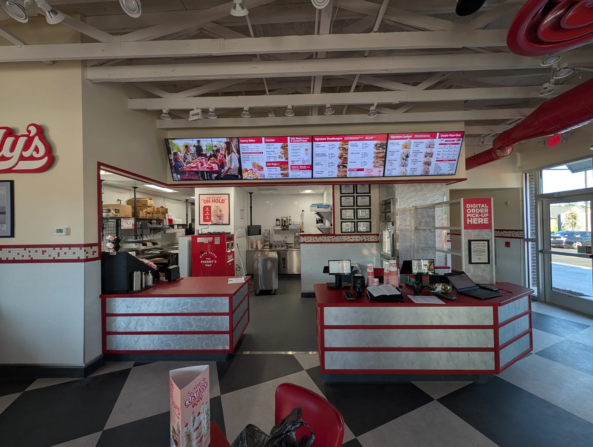 Inside a fast food restaurant, with a digital menu board overhead displaying various food options, a counter with a cash register, and seating with red chairs and checkered flooring.