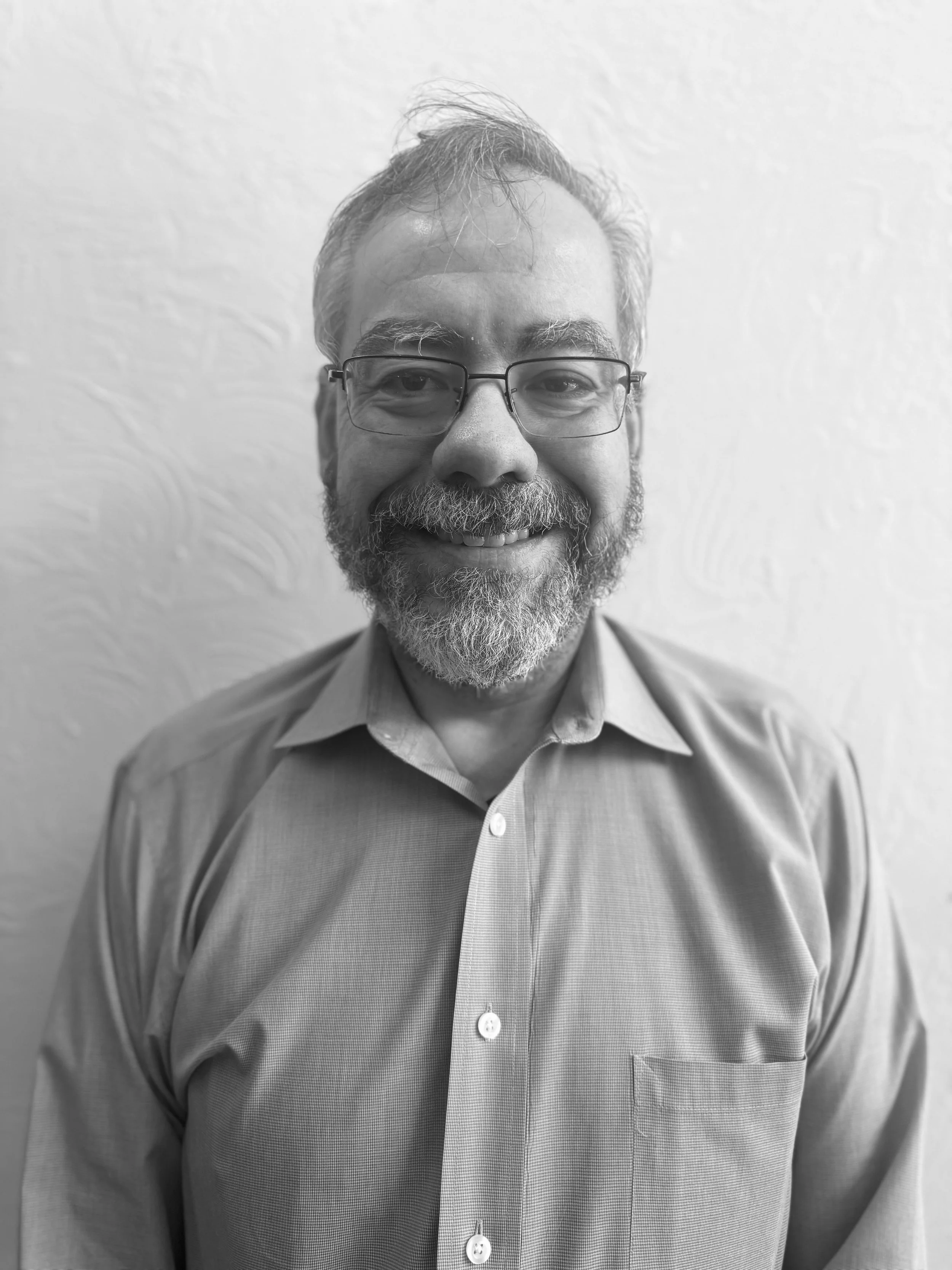 Black and white photo of a middle-aged man with glasses, a beard, and styled hair, smiling and wearing a button-up shirt.