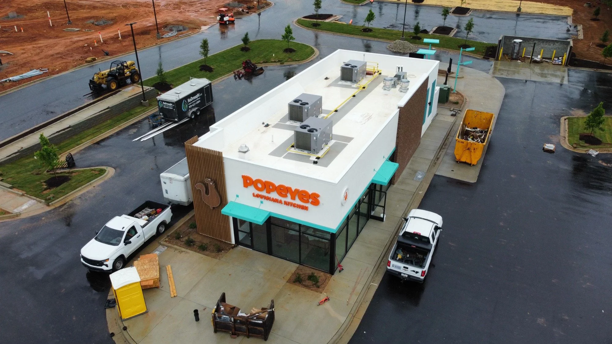 An aerial view of a Popeyes restaurant under construction with a partially paved parking lot, construction vehicles, and equipment, surrounded by wet pavement and small green trees.