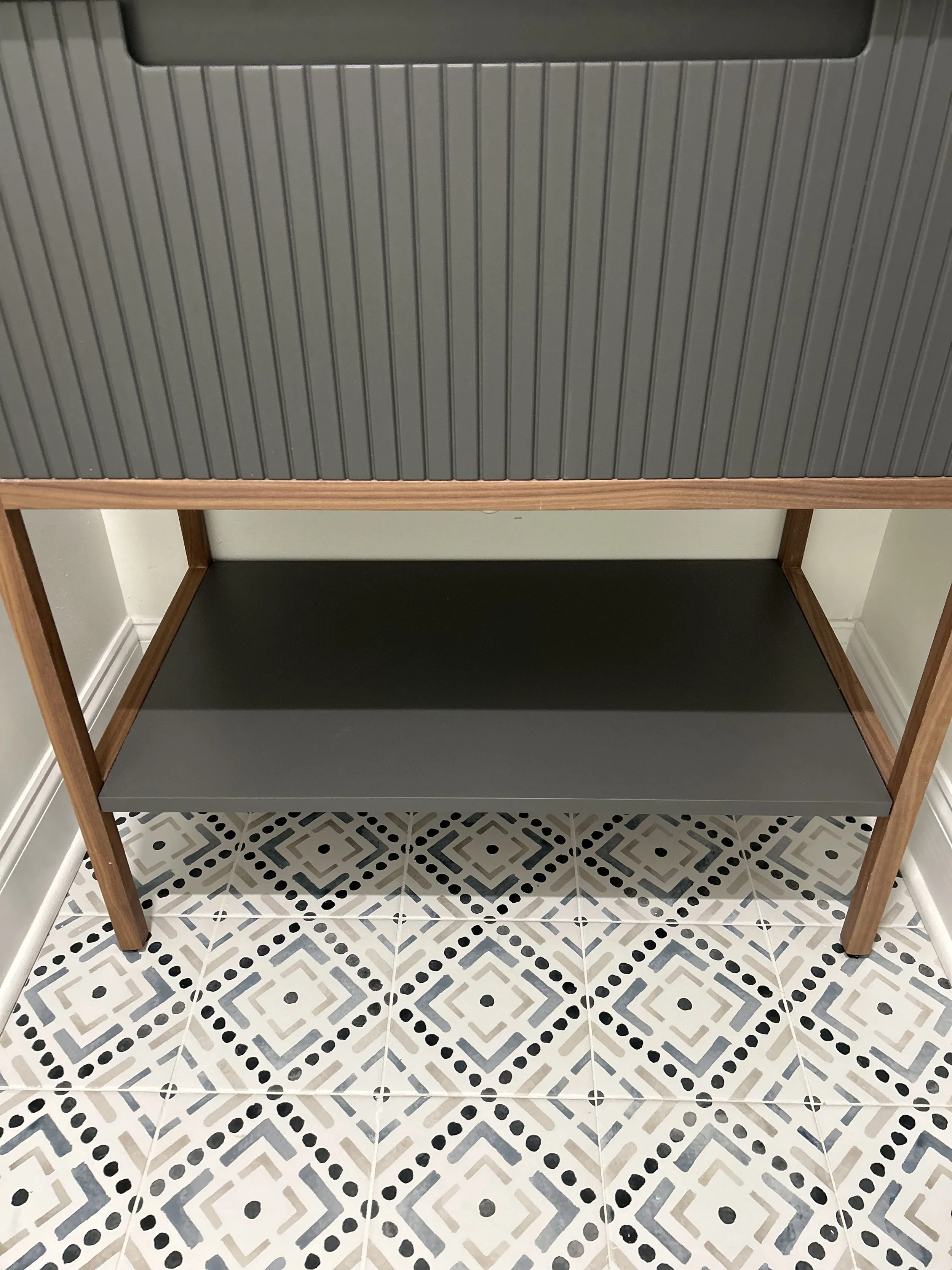 View of a two-tier wooden shelving unit with black shelves, positioned on a patterned tiled floor.