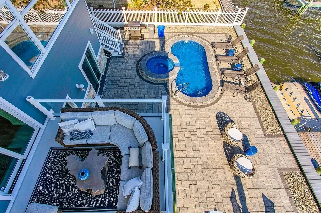 An aerial view of a waterfront backyard showcasing a paved patio with outdoor furniture, a small swimming pool, a hot tub, and a fenced area with lounge chairs and water access.