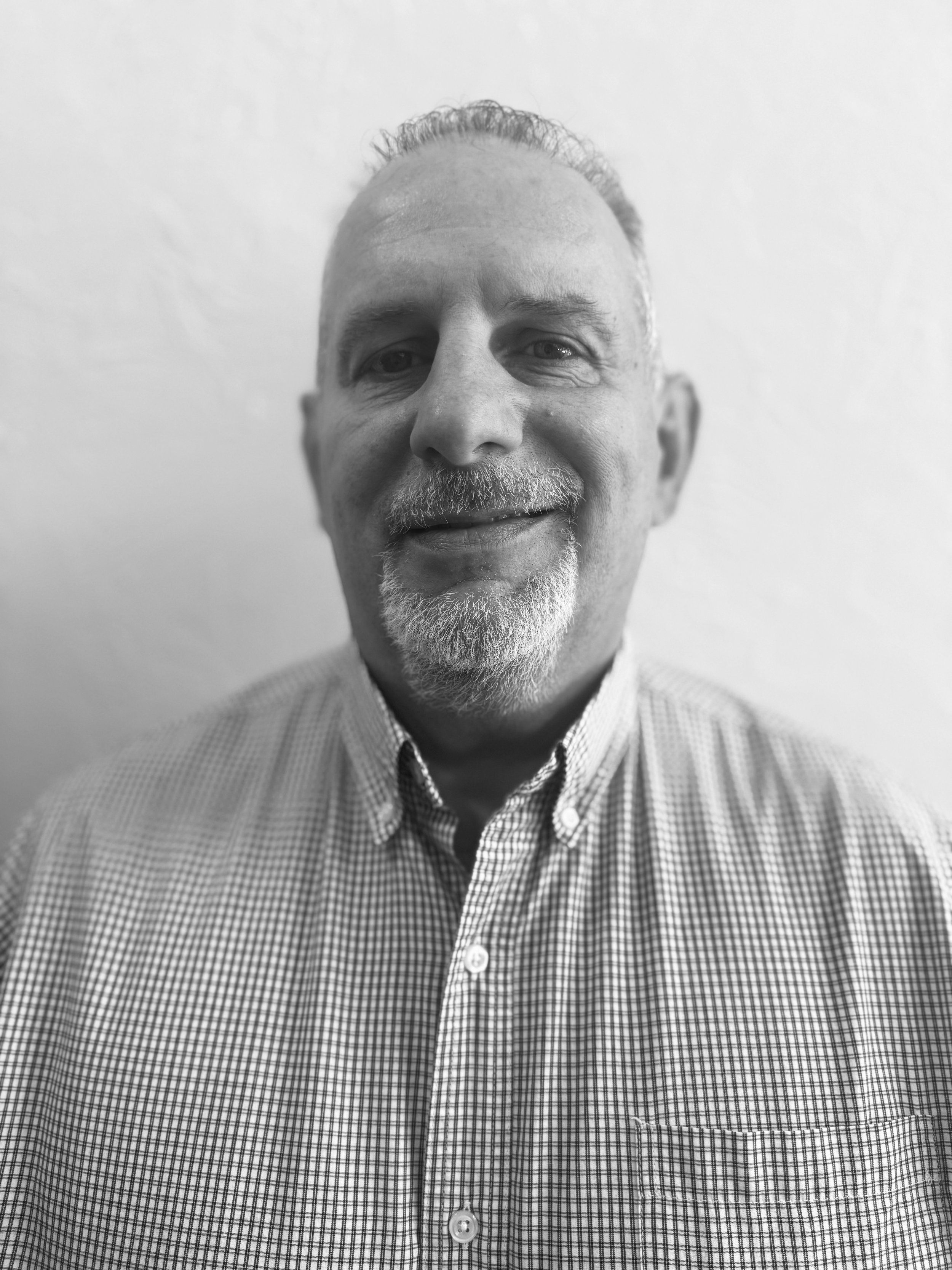 Black and white photo of a middle-aged man with short hair, a goatee, and a checkered shirt, smiling slightly and looking directly at the camera.