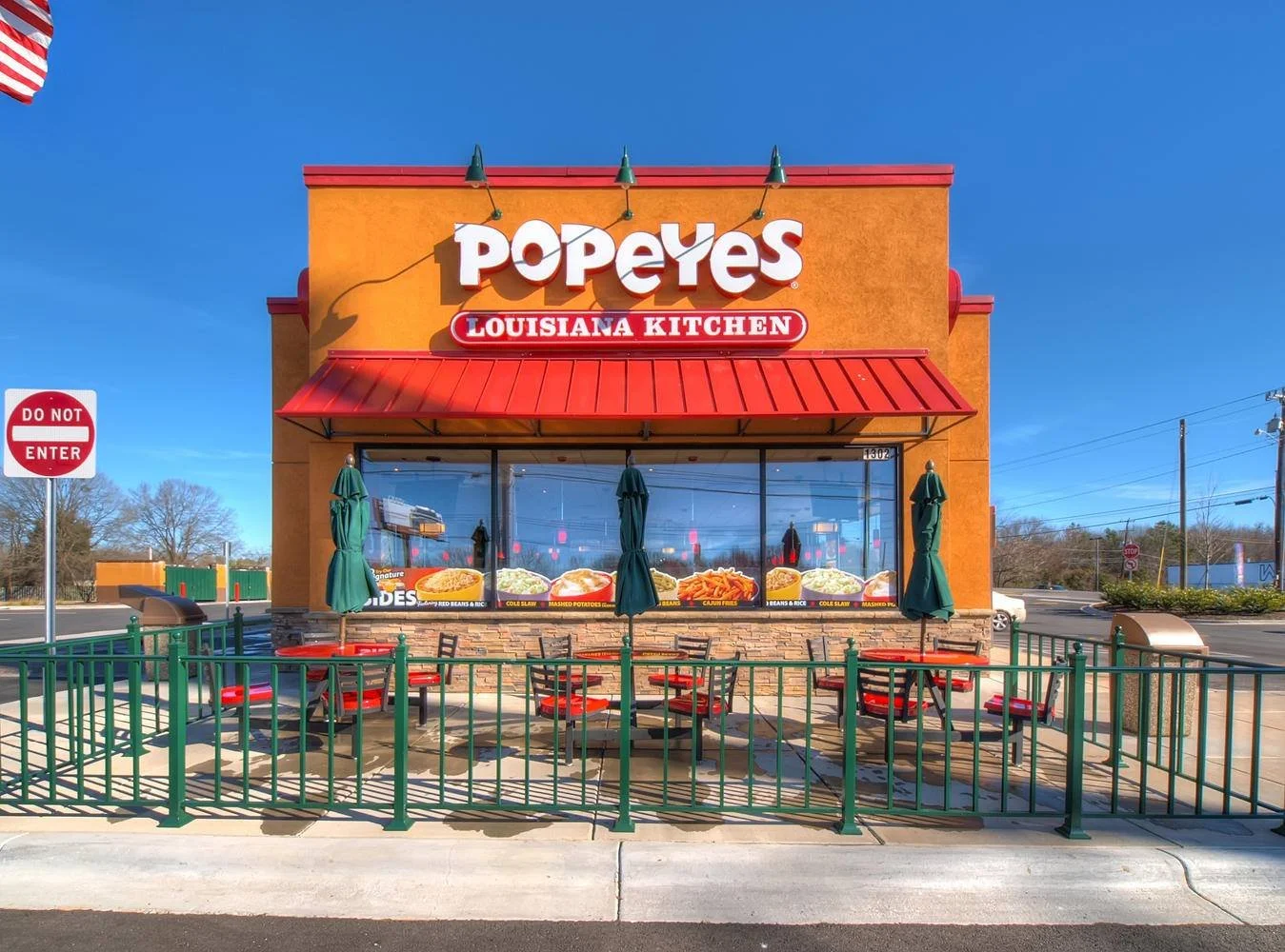 Front view of Popeyes Louisiana Kitchen fast-food restaurant with outdoor seating area, closed green umbrellas, and a clear blue sky.