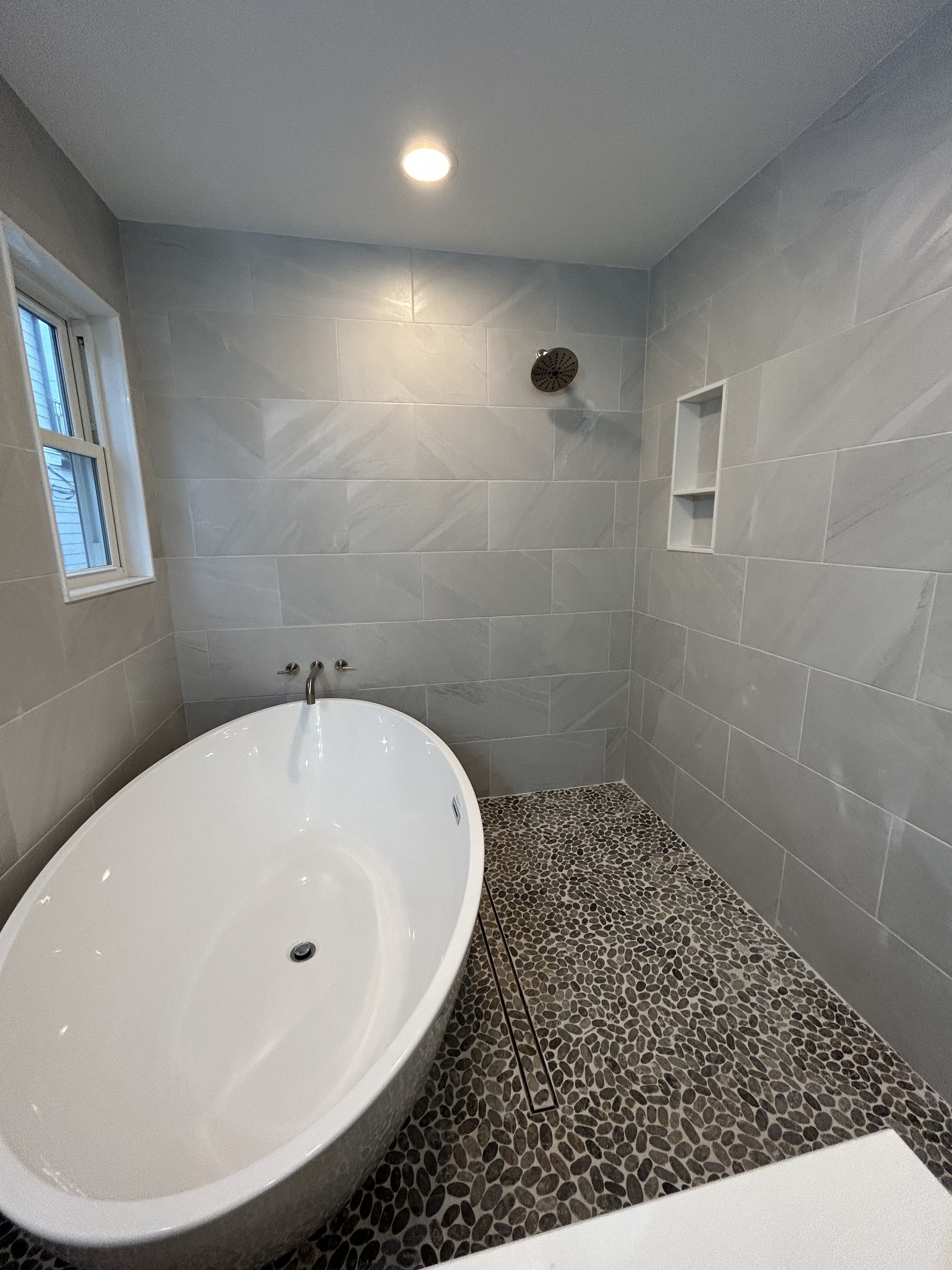 Modern bathroom with a freestanding bathtub, gray tiled walls, a small window, and a pebble-textured floor, with a showerhead visible on the wall.