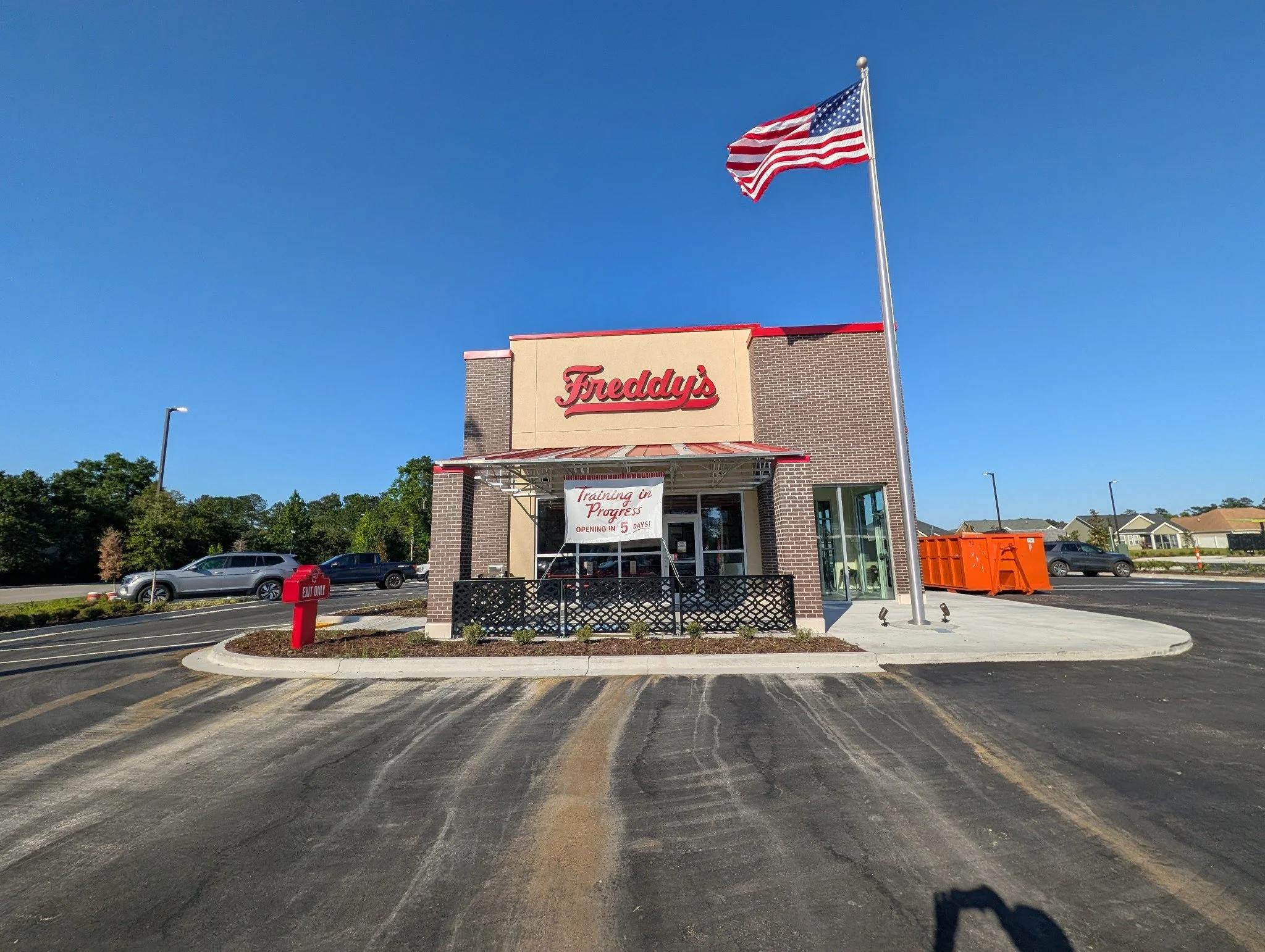 A Freddy's restaurant building with a patriotic American flag on a flagpole in front. The sign on the building reads 'Freddy's' and a banner beneath states 'Training in Progress, Opening in 5 Days!' The parking lot has cars and a red 'Exit Only' sign