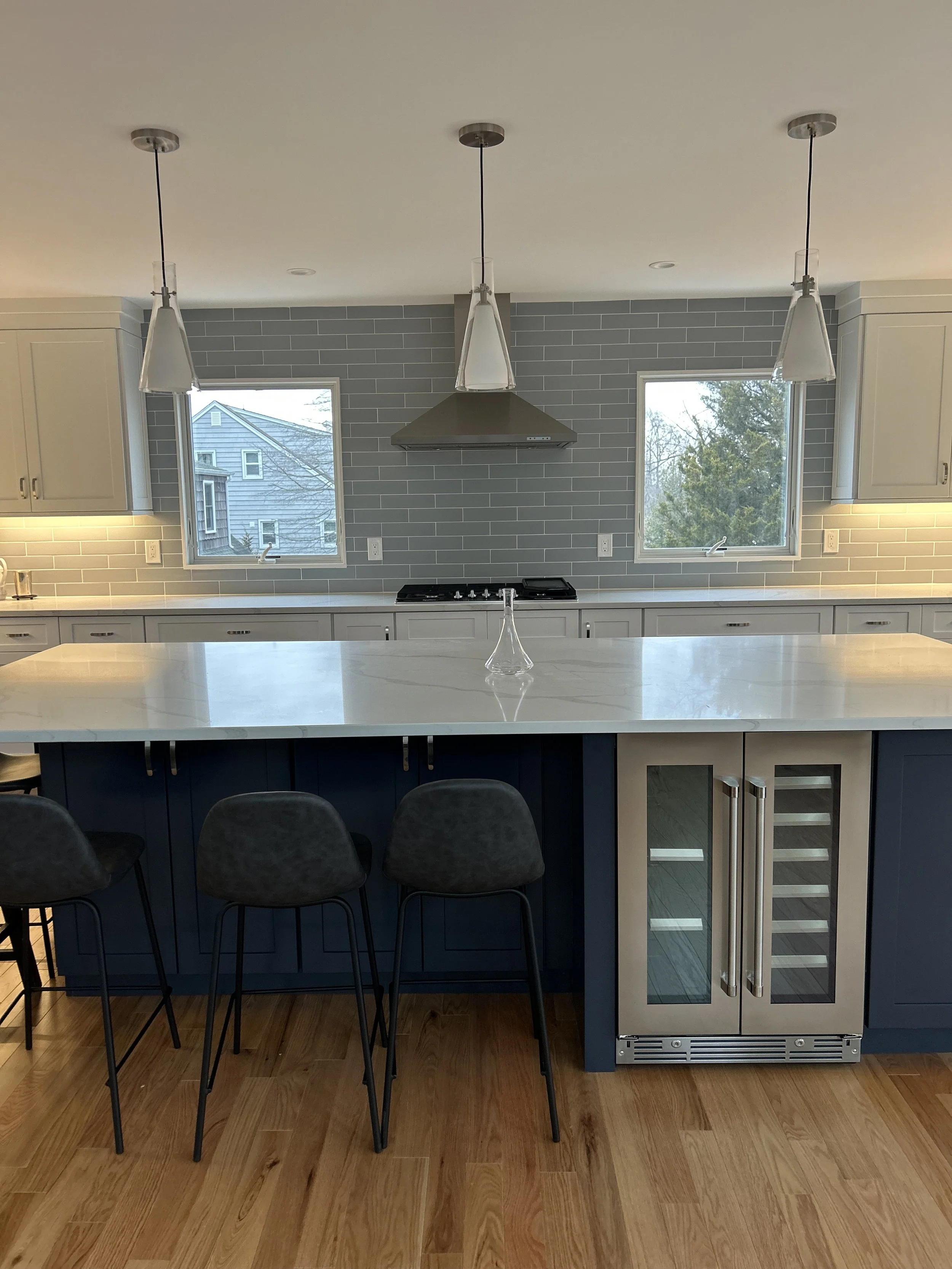 Modern kitchen with white countertops, gray subway tile backsplash, three pendant lights, two windows, stove, and bar stools with black cushions.