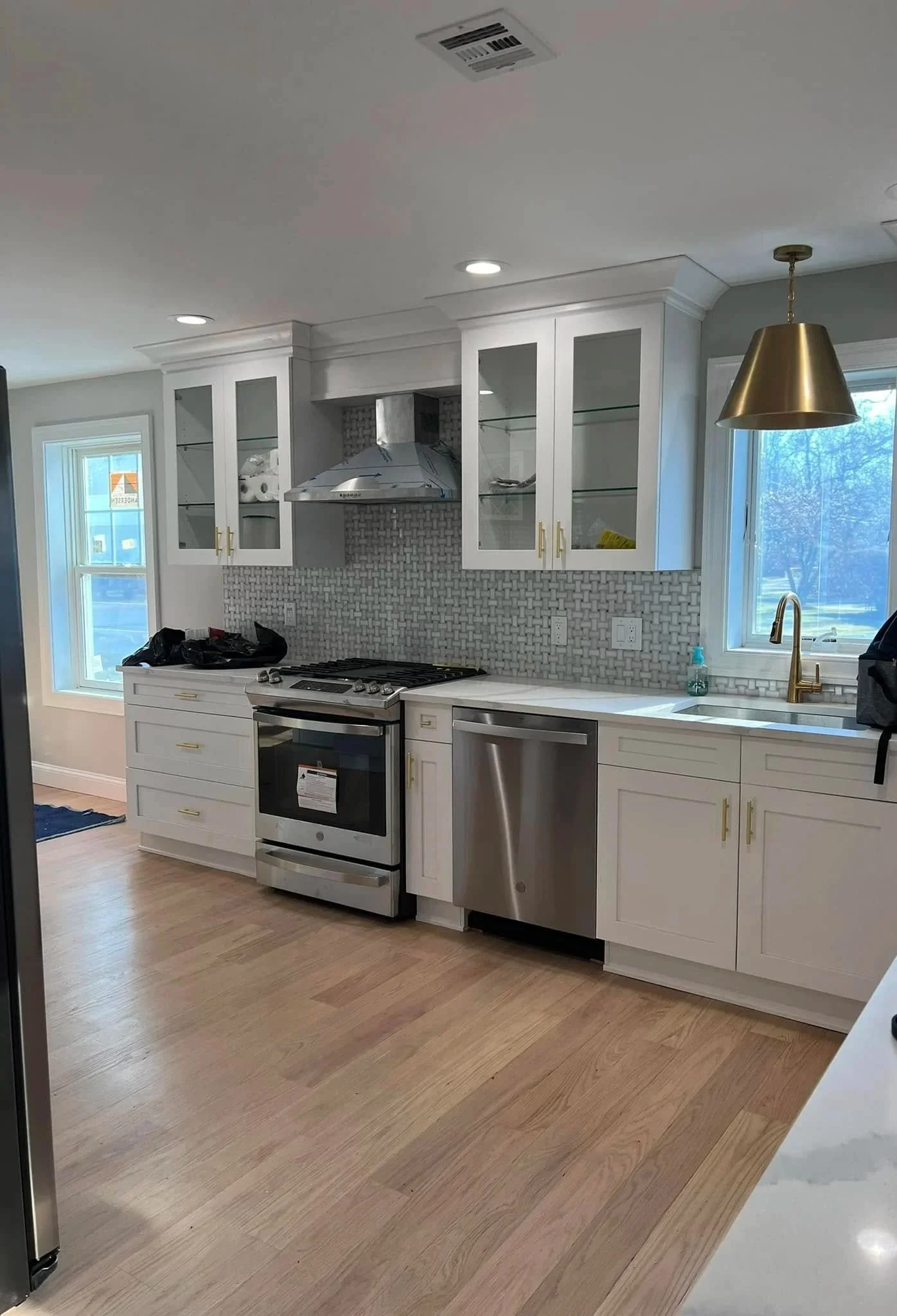 Modern kitchen with white cabinets, stainless steel oven and dishwasher, glass-front upper cabinets, a countertop with a small bottle of hand soap, a gold hanging light, window showing outdoor trees, and a tiled backsplash.