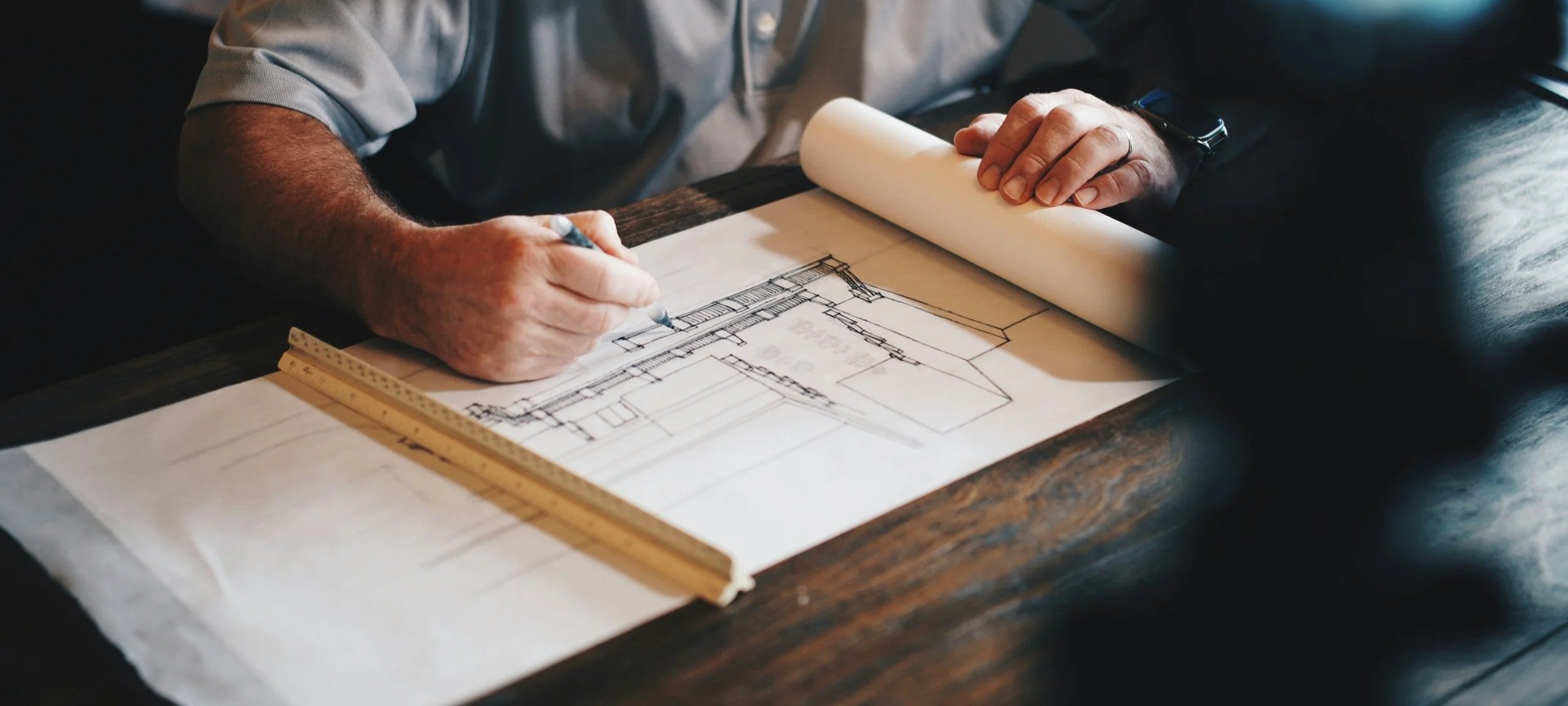 A person drawing architectural blueprints on paper at a wooden table.