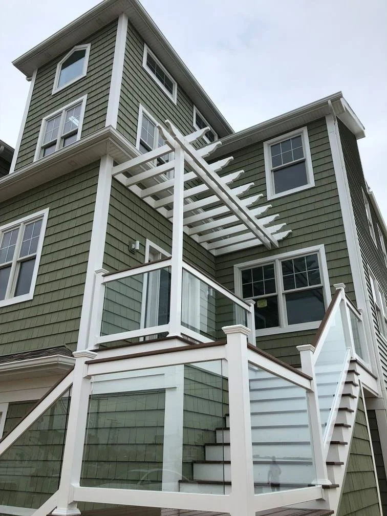 Multi-story house with green siding and white trim, featuring glass-paneled balcony railings and an external staircase leading to a rooftop deck under construction.