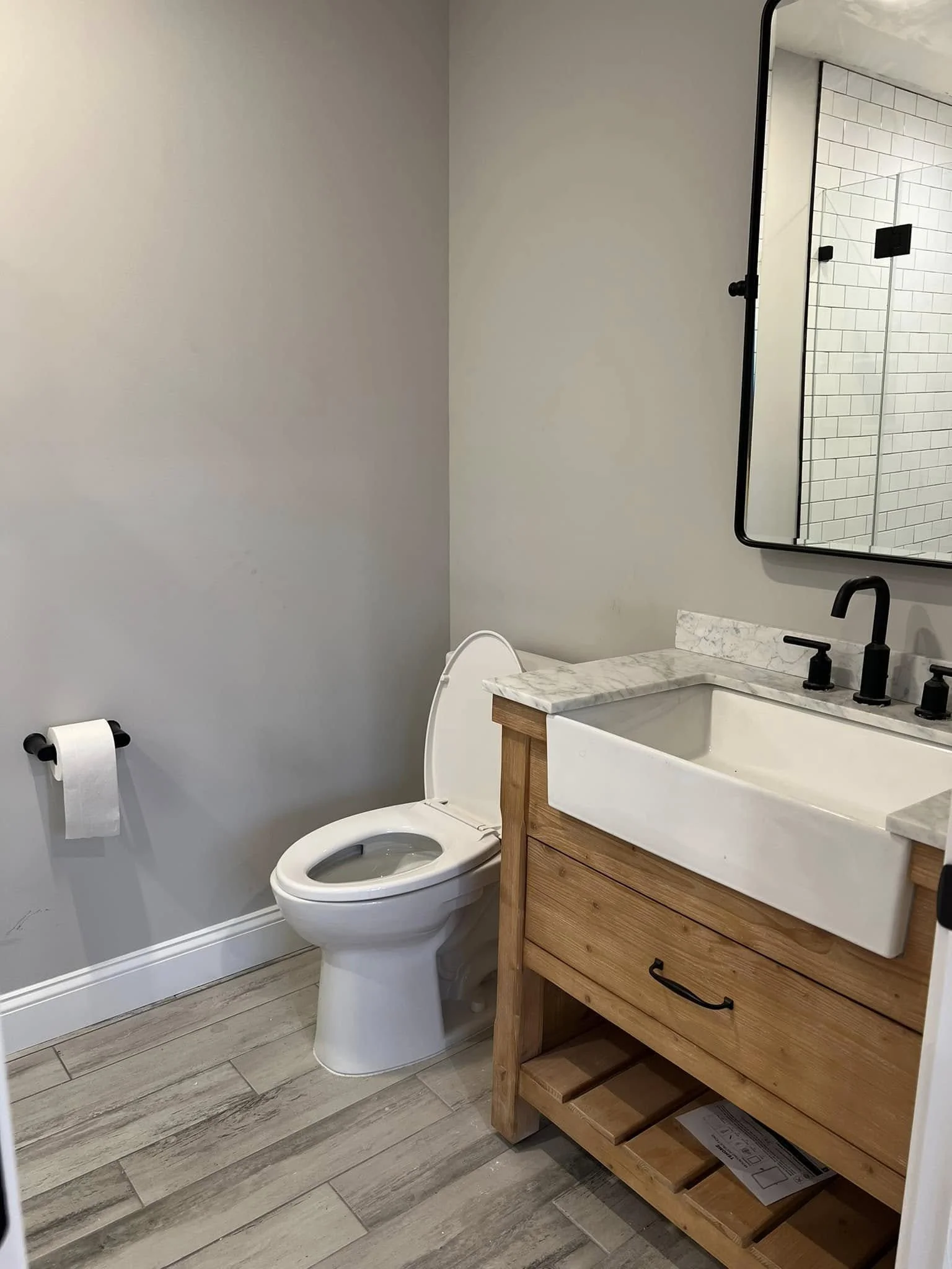 A bathroom with a white toilet, a wooden vanity with a white sink, black faucet and fixtures, a mirror, and a gray wall. The bathroom has wood-look tile flooring and a white tiled shower area with a black showerhead and controls visible in the reflec