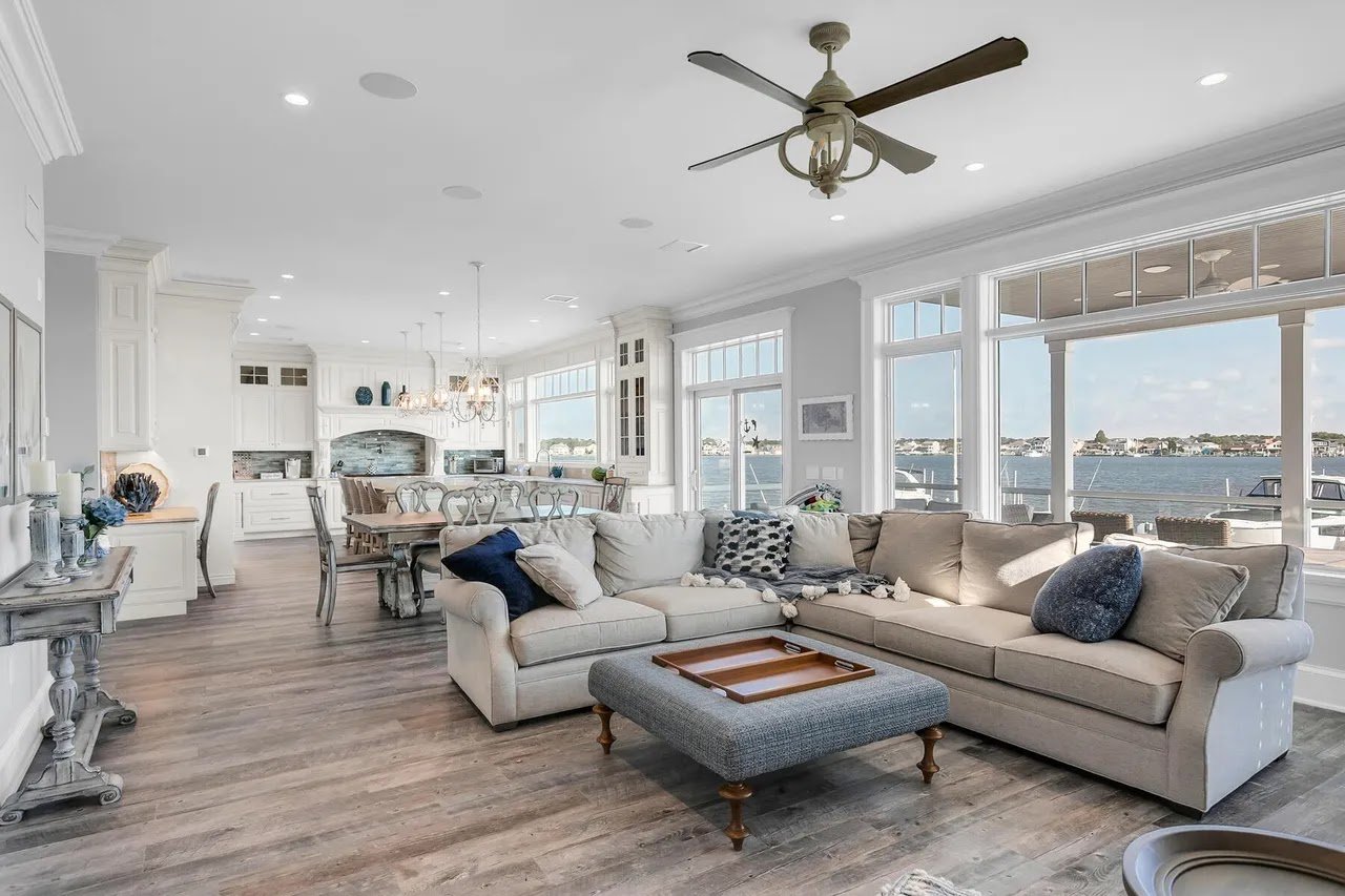 Open concept living room and kitchen with large windows overlooking the water, light-colored furniture, and wood flooring.