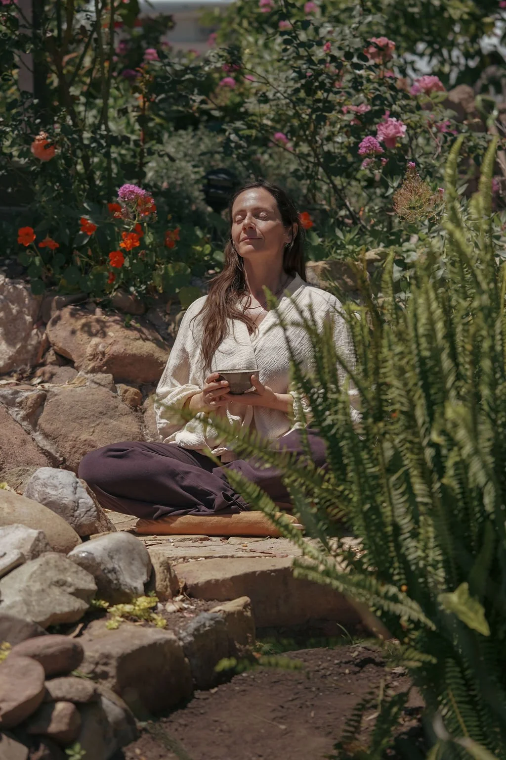 A woman with long brown hair, sitting cross-legged on a stone pathway in a garden, holding a small bowl with eyes closed and a peaceful expression, surrounded by various flowering plants and rocks.