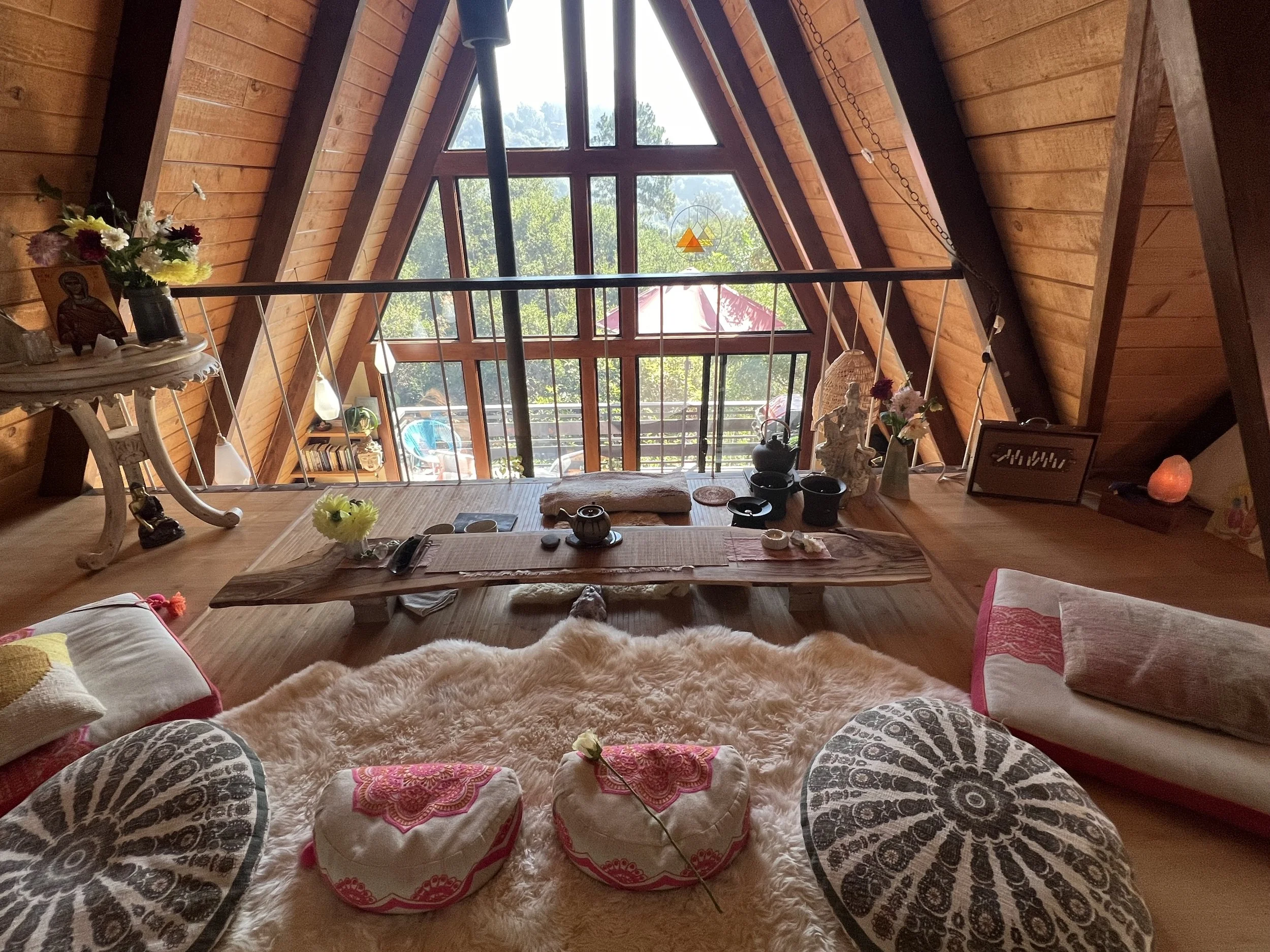 The inside of an A-frame cabin with a high triangular window, wooden walls, and a cozy seating area with patterned cushions and blankets. Decor includes flowers, sculptures, and various small objects on a low table.