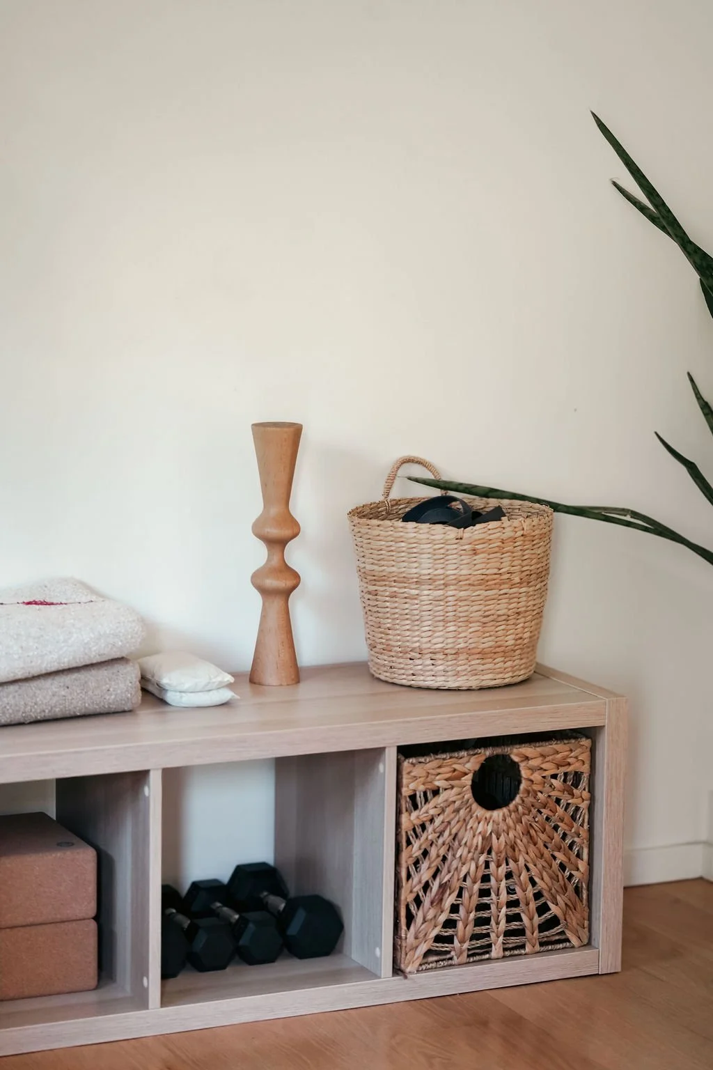 A wooden shelf with folded towels and pillows on the left, a wooden decorative candle holder, and a woven basket with headphones on top, placed against a plain wall with a tall green plant on the right.