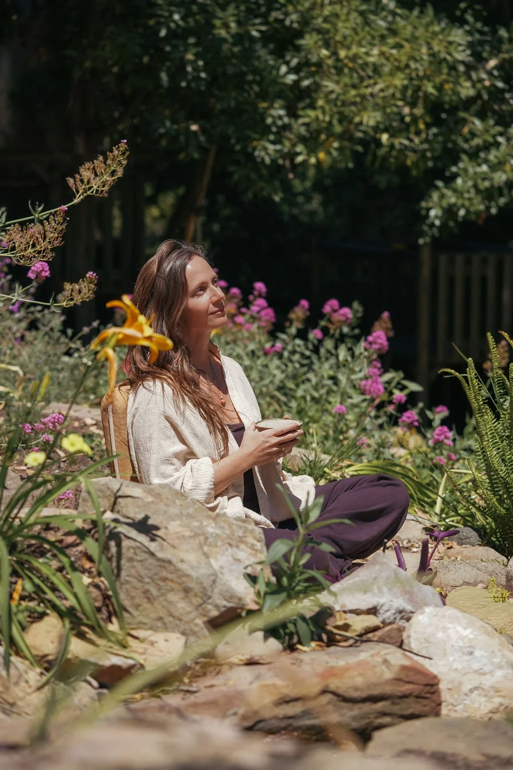 A woman with long brown hair sits on rocks in a garden filled with colorful flowers, holding a cup and enjoying sunlight.