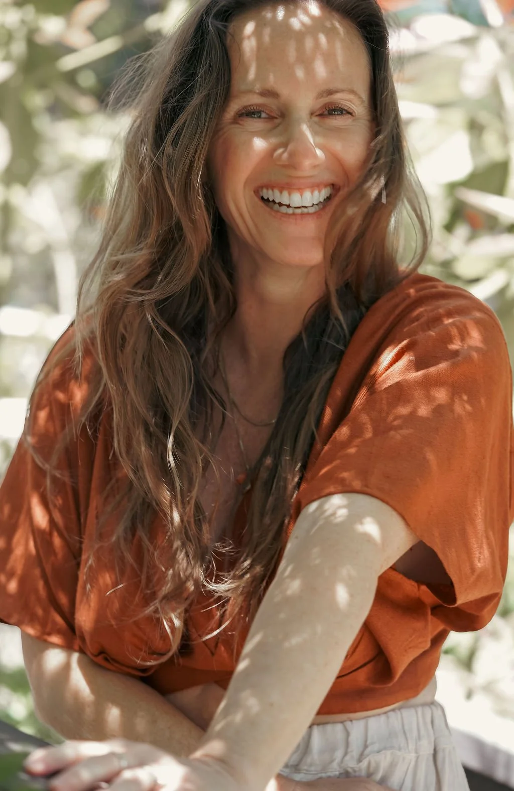 A woman with long brown hair and light skin is smiling outdoors with sunlight filtering through leaves above her. She is wearing an orange shirt and light-colored pants.