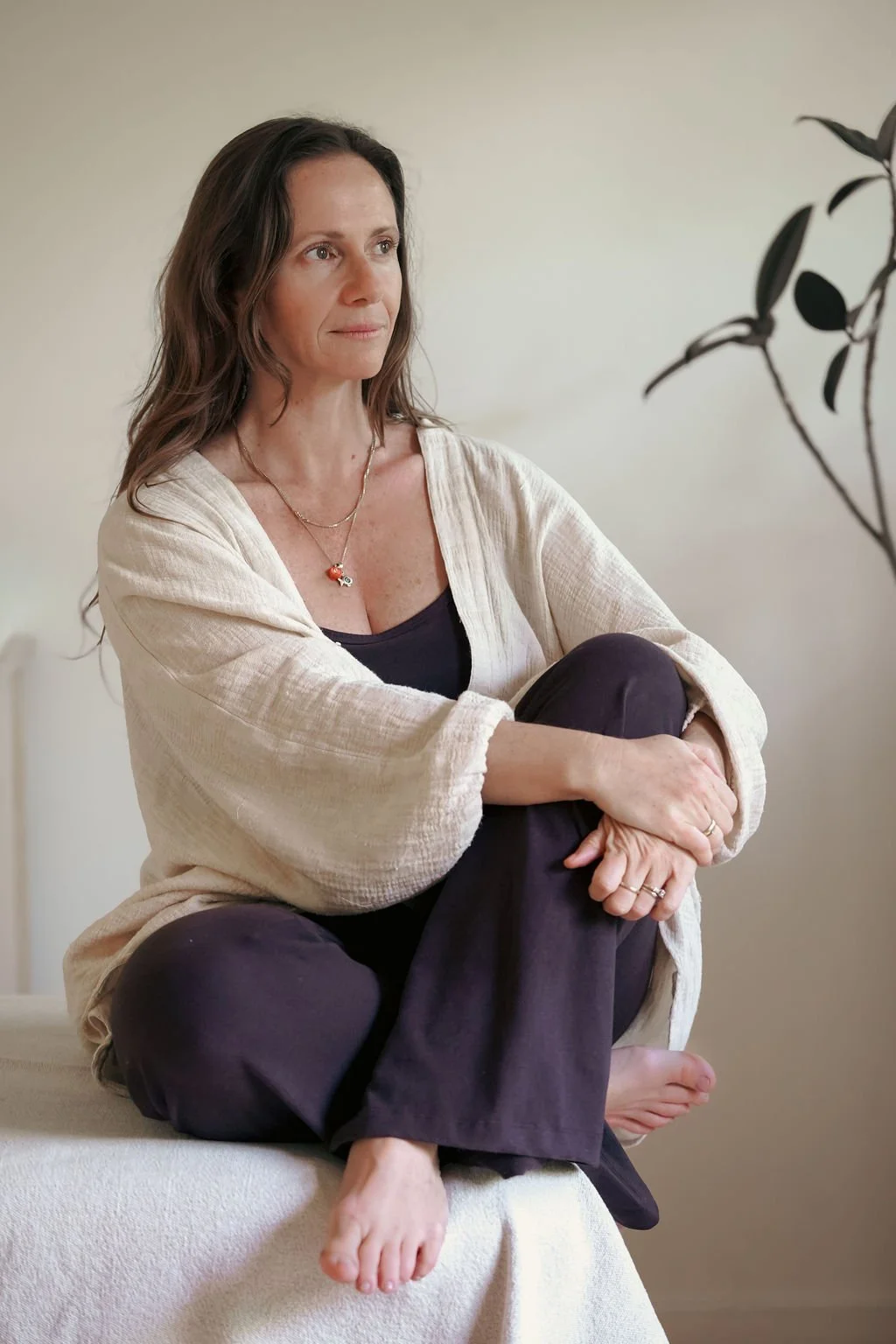 Barbara Stamis, breathwork and bodyworker,  with shoulder-length brown hair sitting on a white surface, wearing a cream-colored cardigan over a black top, with a necklace, black pants, and barefoot, gazing thoughtfully to her left.