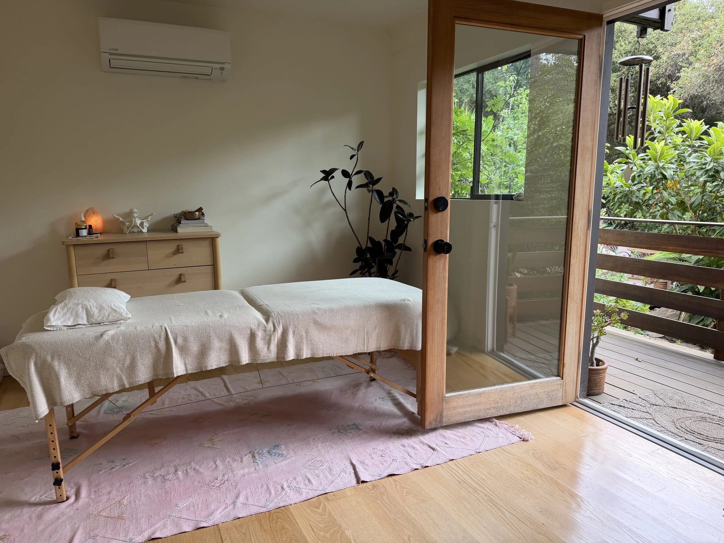 A bedroom with a massage table, a wooden chest of drawers with small items on top, a plant, and glass sliding doors leading to a porch with greenery outside.