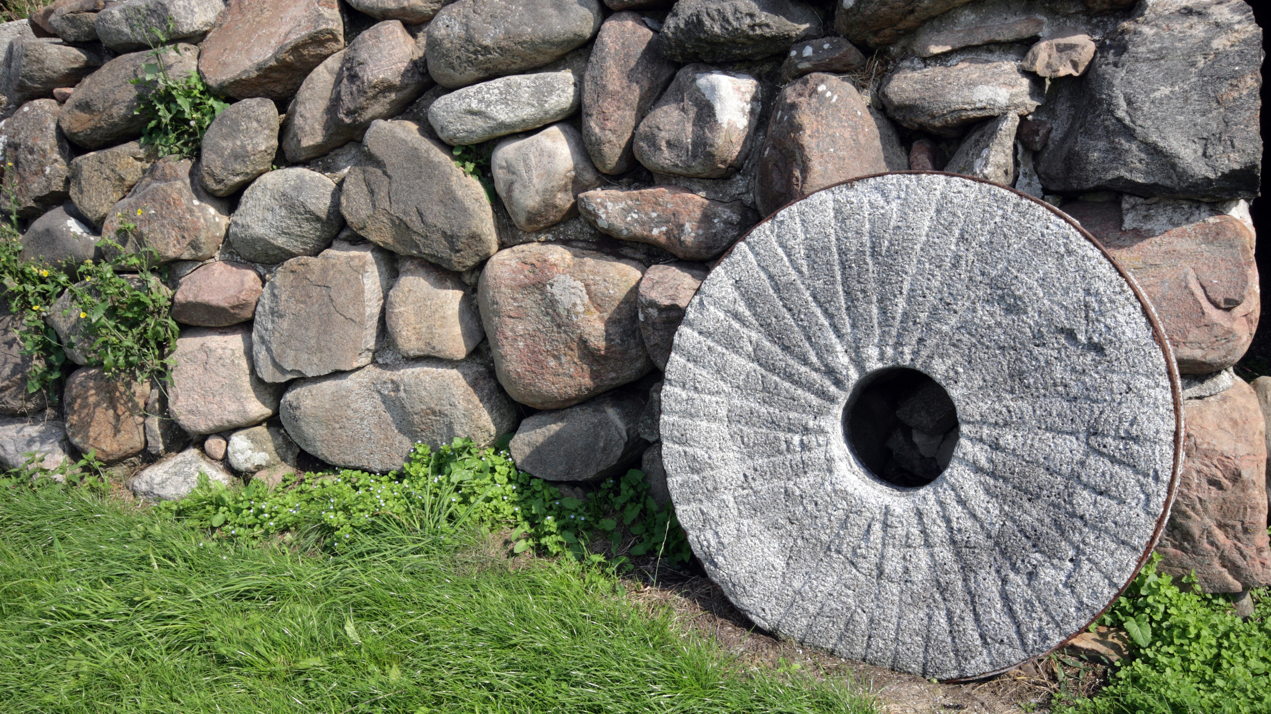 A stone wall with a large, circular millstone resting against it. The wall is composed of various-sized roughly shaped stones, and there is green grass and small plants growing at the base of the wall.