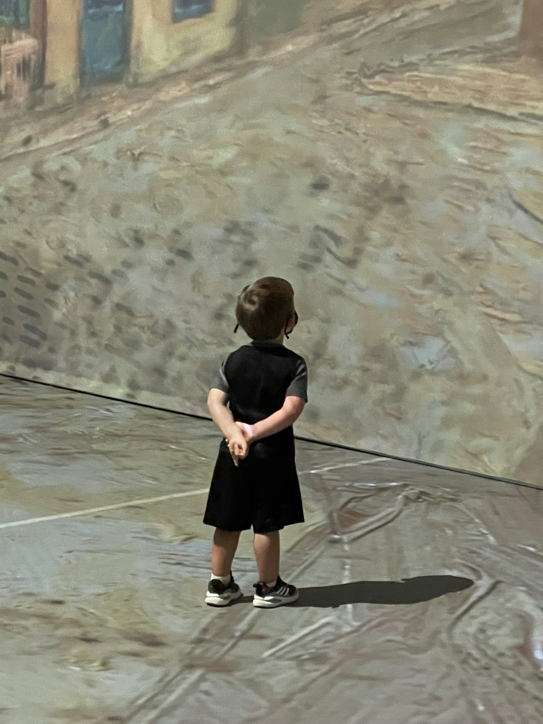 A young person with short hair, wearing a black shirt, black shorts, and sneakers, stands with hands clasped behind back, looking at a large textured abstract painting or mural in an art gallery or museum.