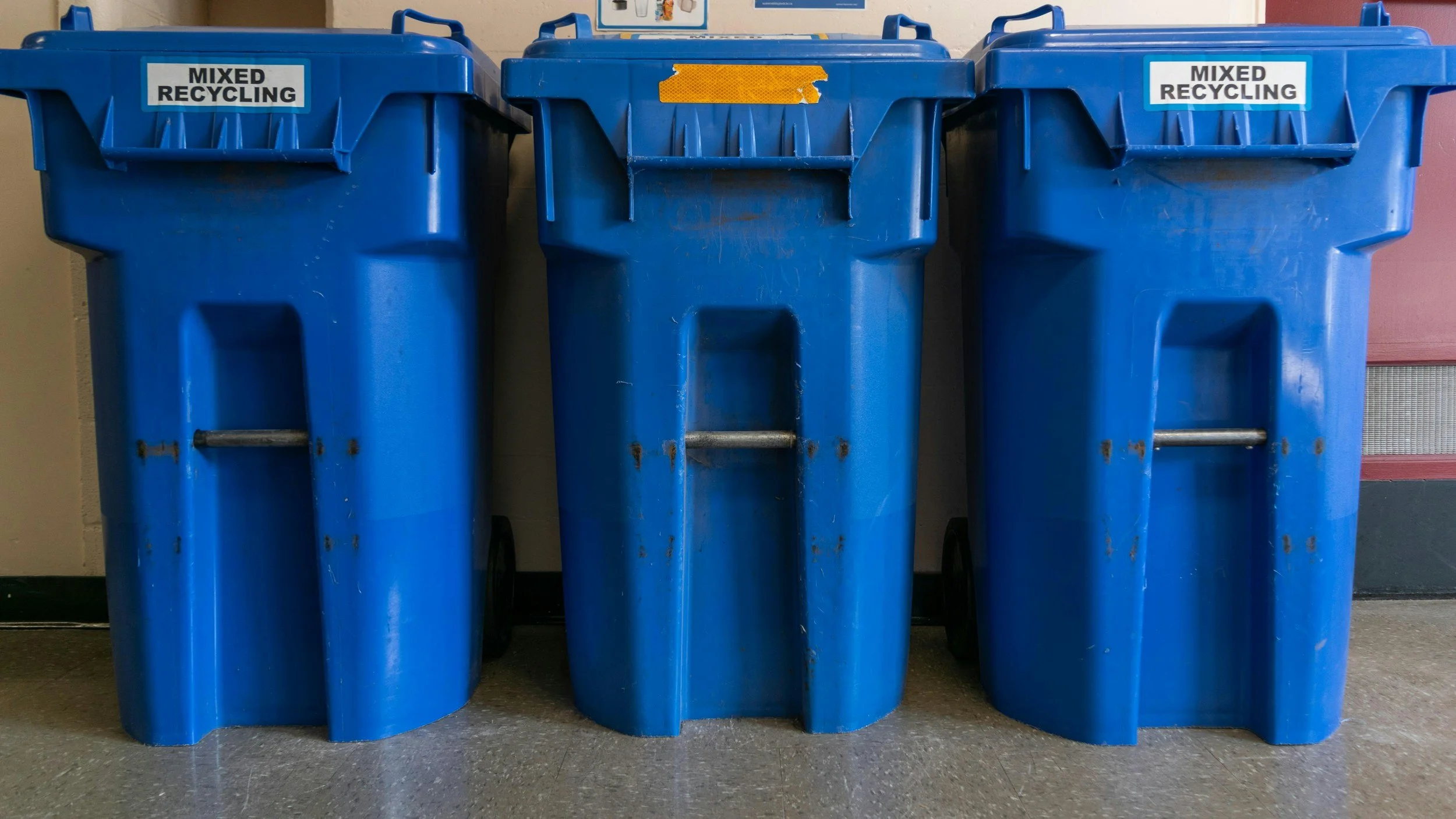 Three blue recycling bins labeled 'Mixed Recycling' lined up against a wall.