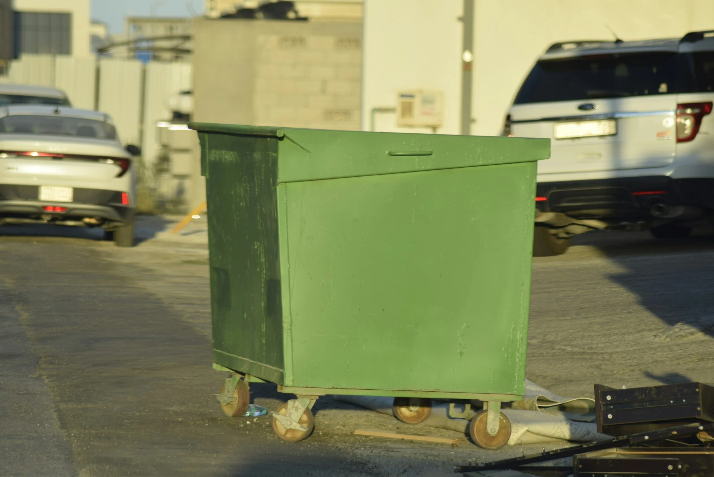 A green trash cart on wheels situated in a parking lot, with several parked cars visible in the background.