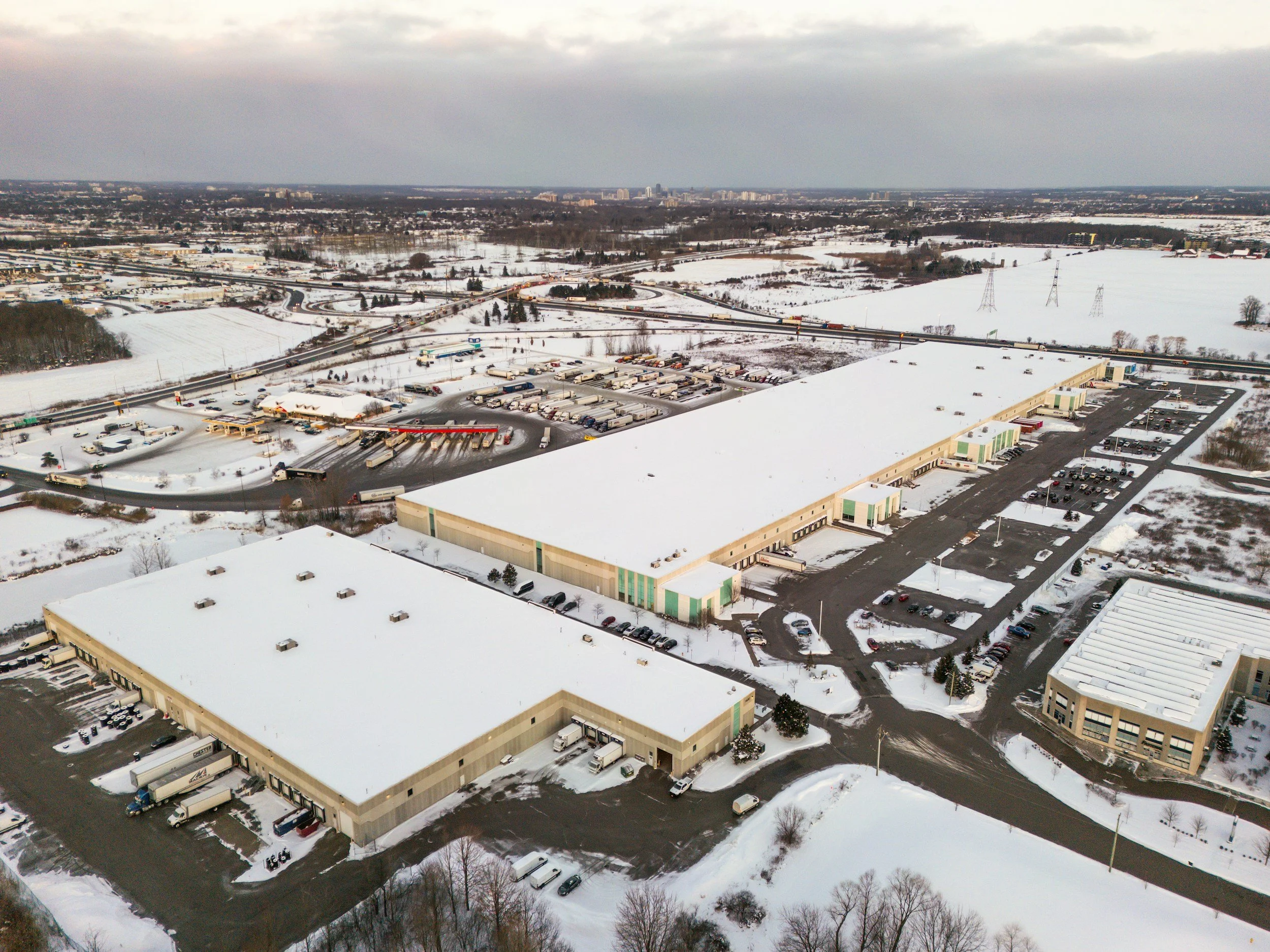 Aerial view of a snowy industrial and commercial area with large buildings, parking lots, and a highway with truck traffic on a winter day.
