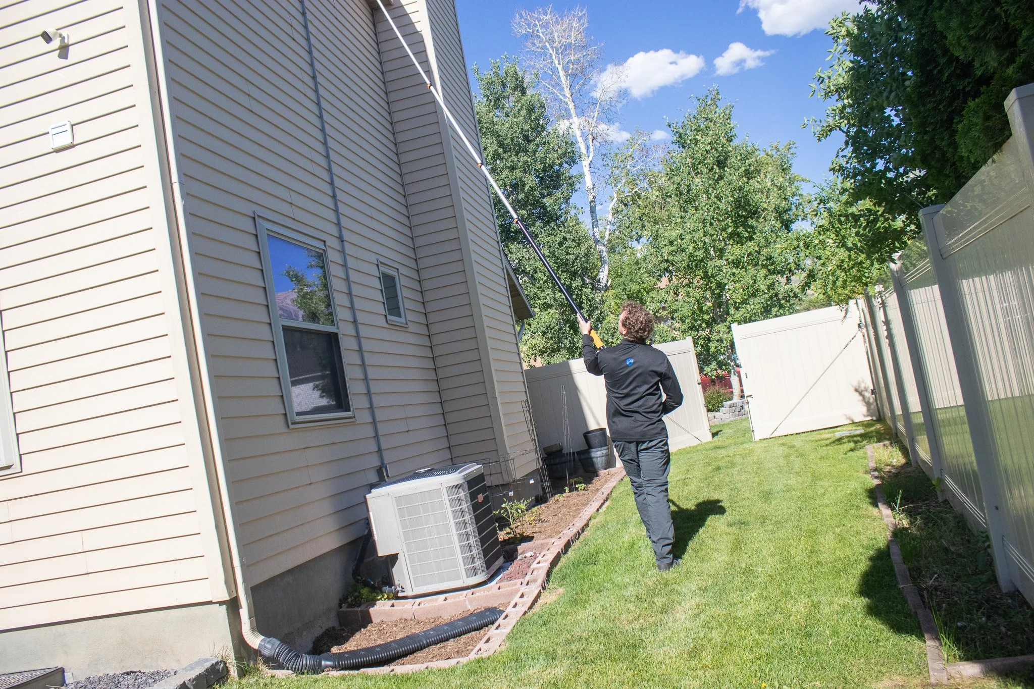 A person using an extendable pole to clean or inspect the side of a two-story house with beige horizontal siding, windows, and air conditioning units in the backyard on a sunny day with a blue sky and green trees.