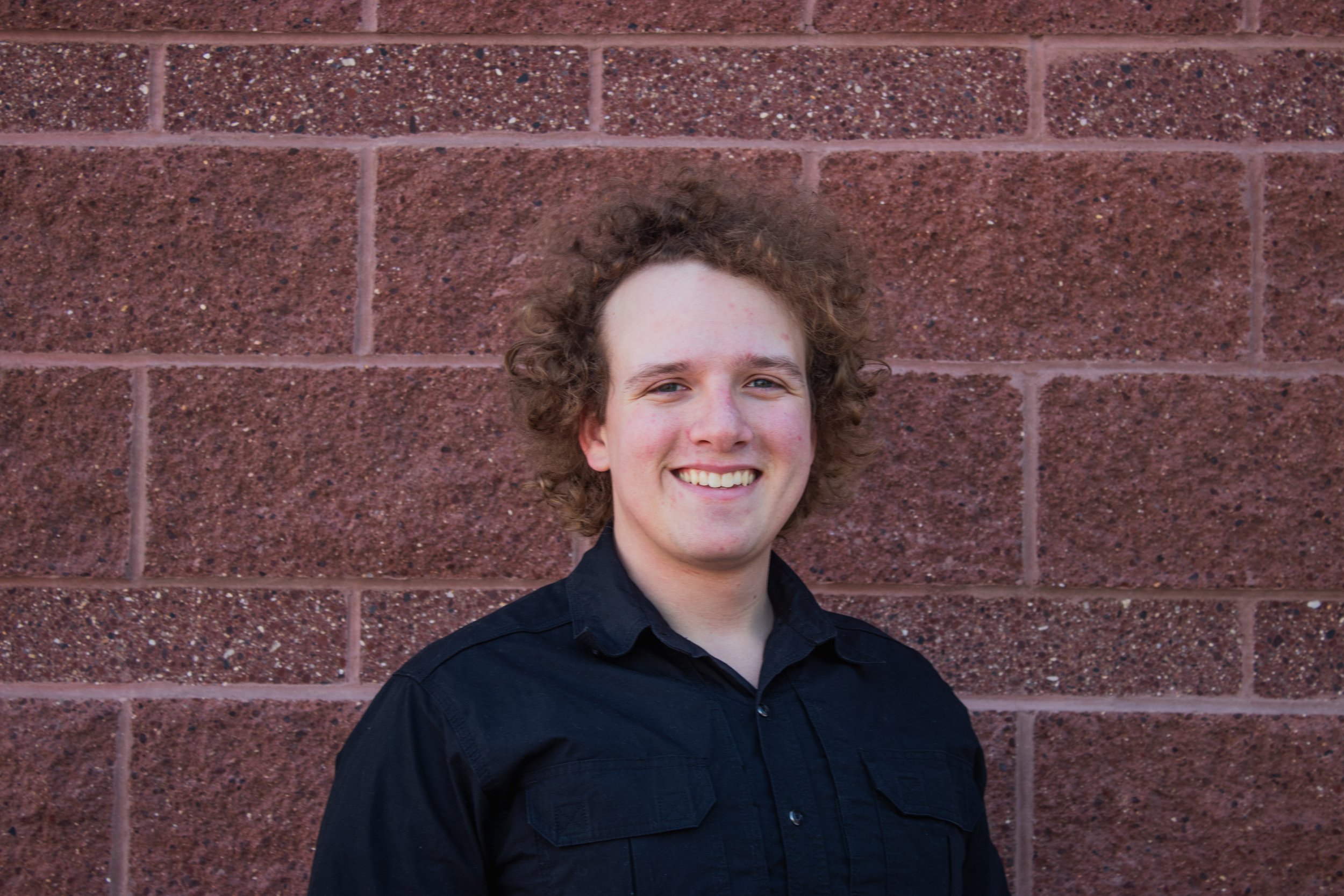 A young man with curly hair smiling, wearing a black shirt, standing in front of a red brick wall.