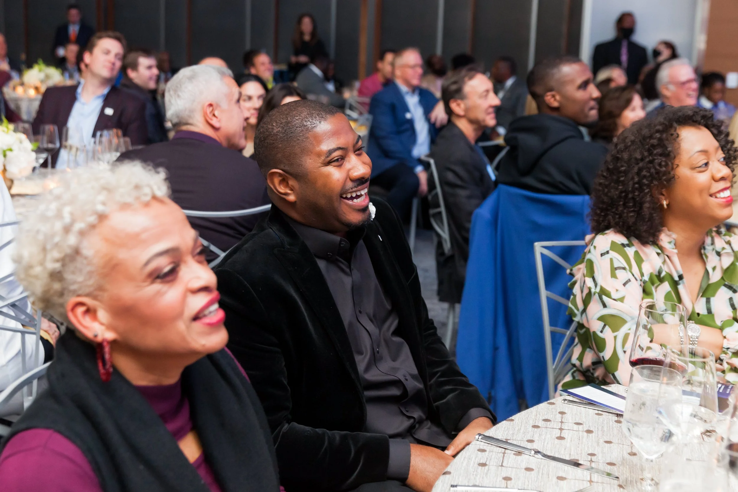 A diverse group of people sitting at a dining table, smiling and enjoying a formal event, celebration or speech.