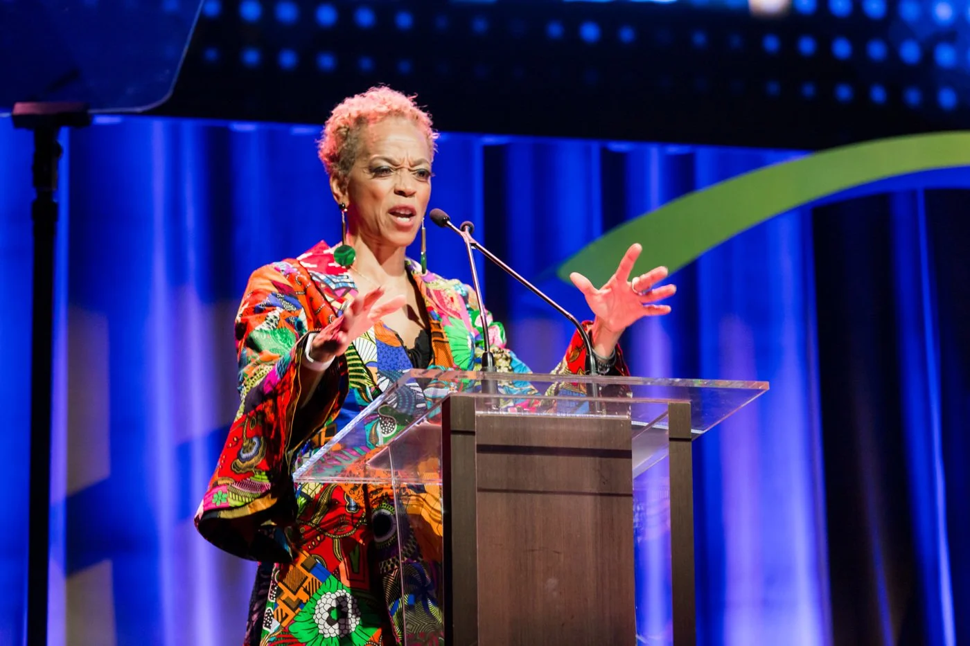 A woman with short curly hair and vibrant colorful clothing speaking at a podium on a stage with blue curtains in the background.