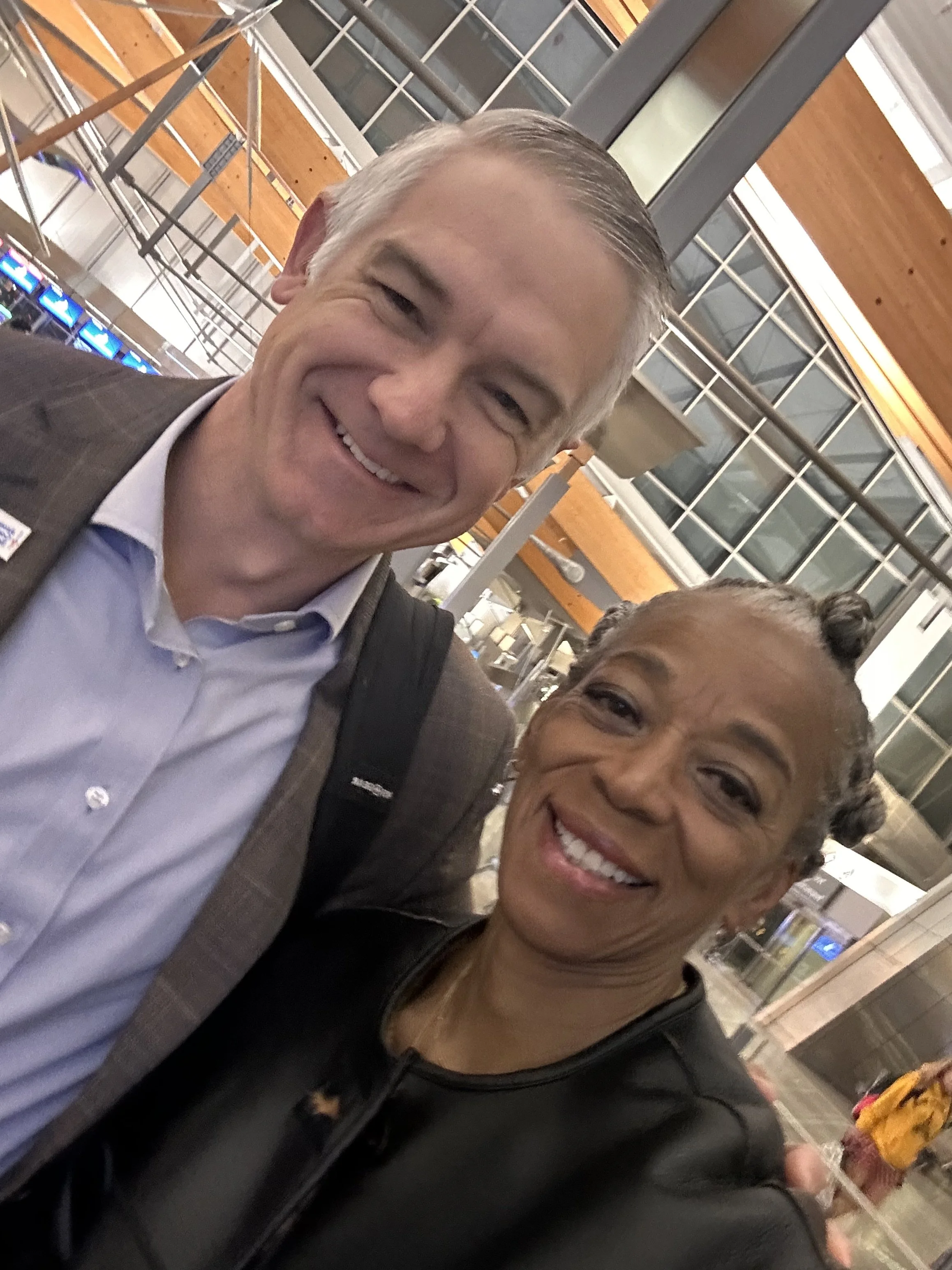 A smiling elderly man in a suit and a smiling woman with curly hair in a black top, taking a selfie together at an airport.