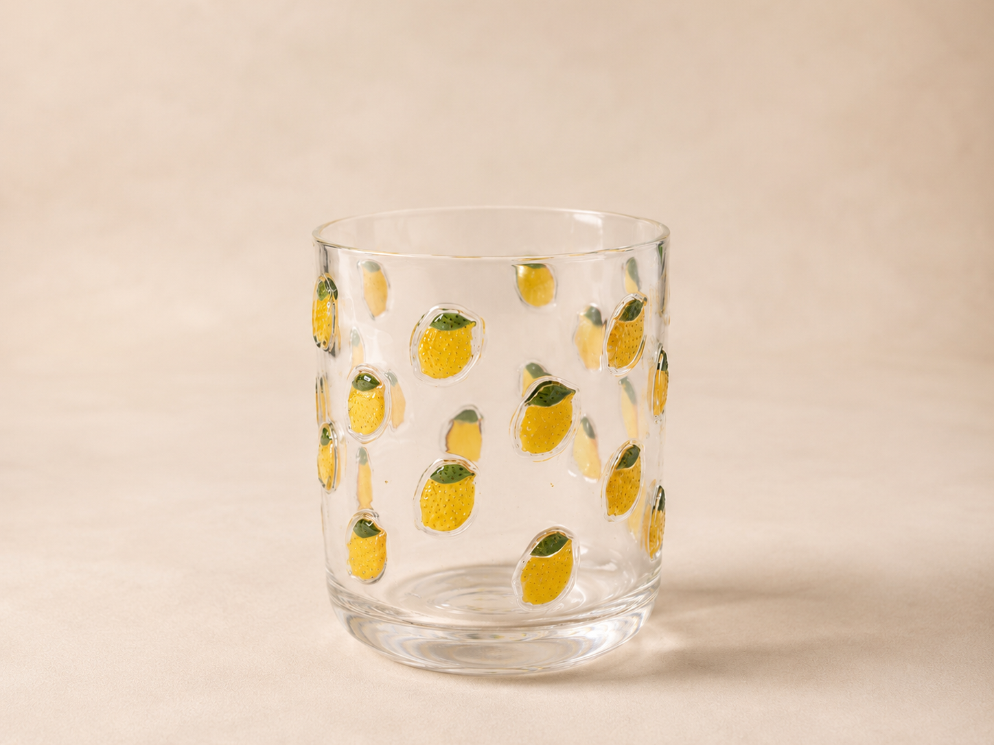 A clear glass tumbler decorated with yellow lemon designs with green leaves, placed on a light beige surface against a neutral background.