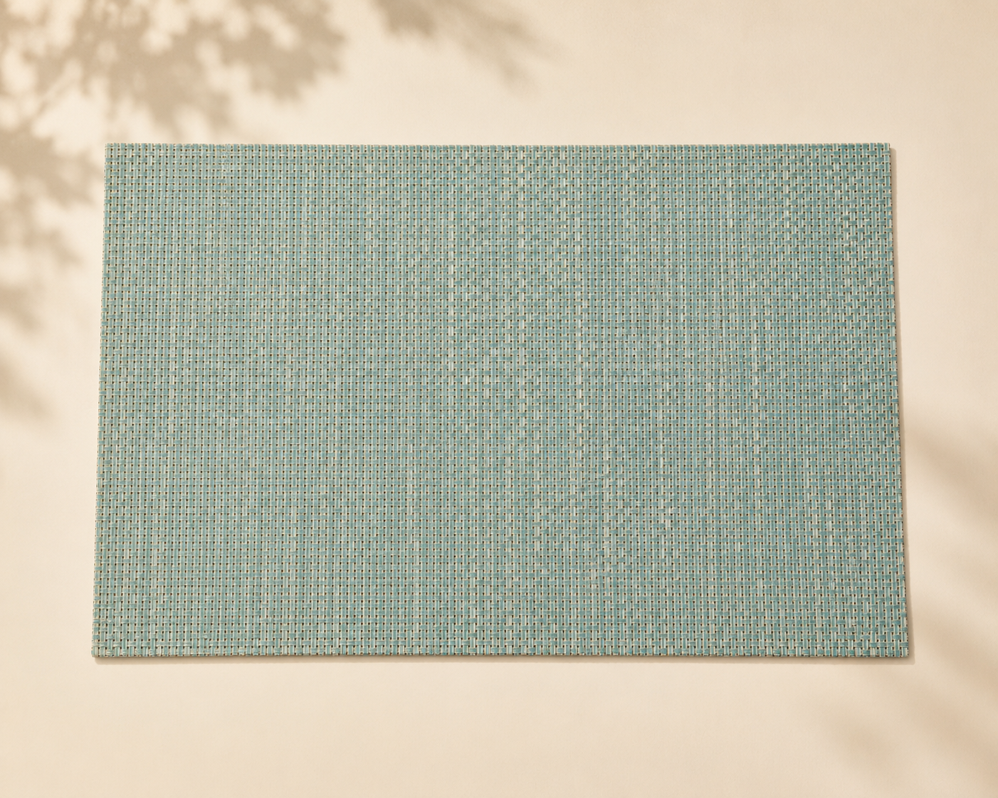 A rectangular light blue woven placemat on a white surface, with blurred shadows of leaves in the background.
